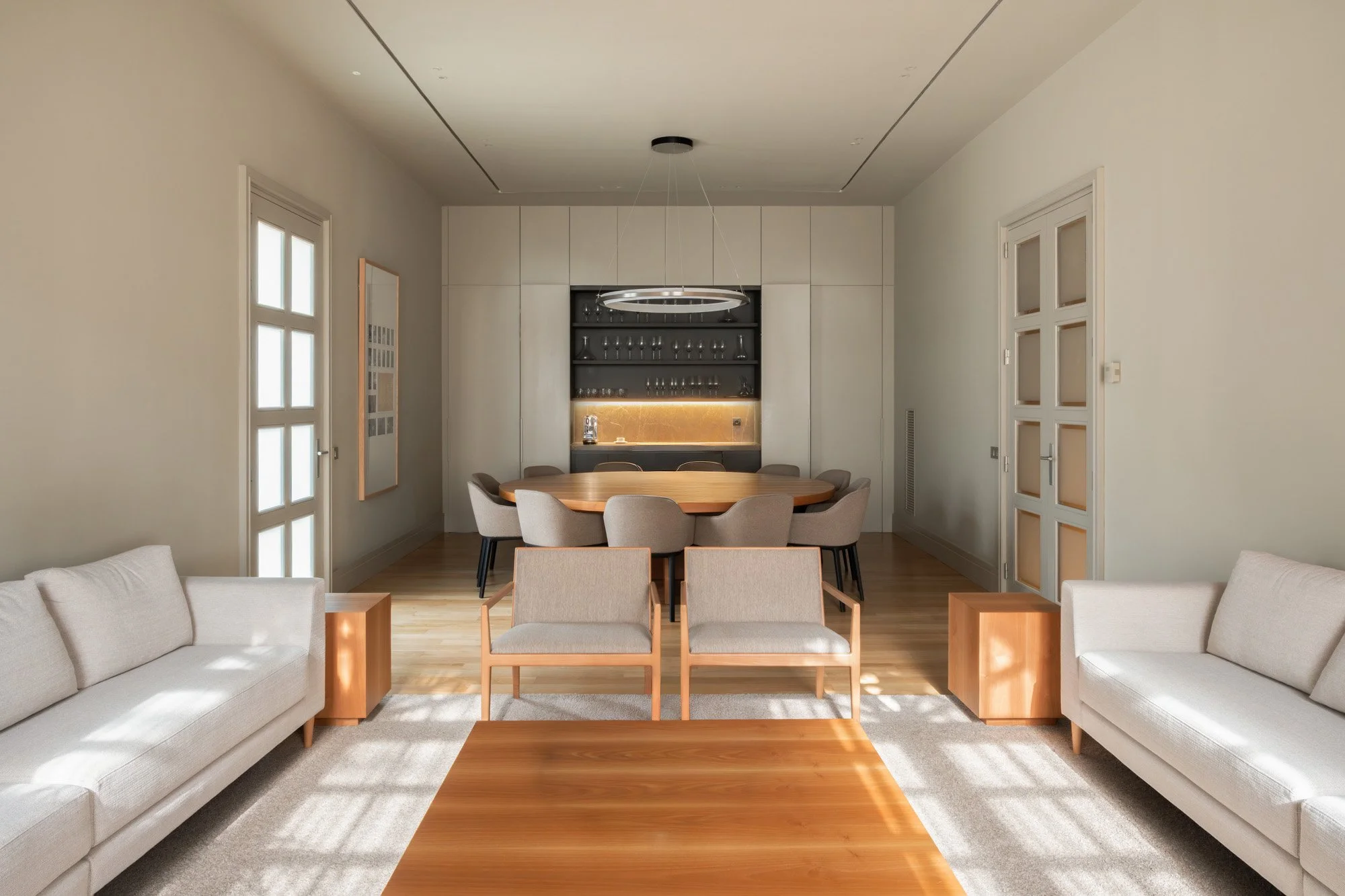 Sala de estar moderna con sillones blancos, mesa de madera, y espacio para comer con una mesa ovalada y sillas beige. En el fondo, bar con cristalería y armarios blancos y negros.