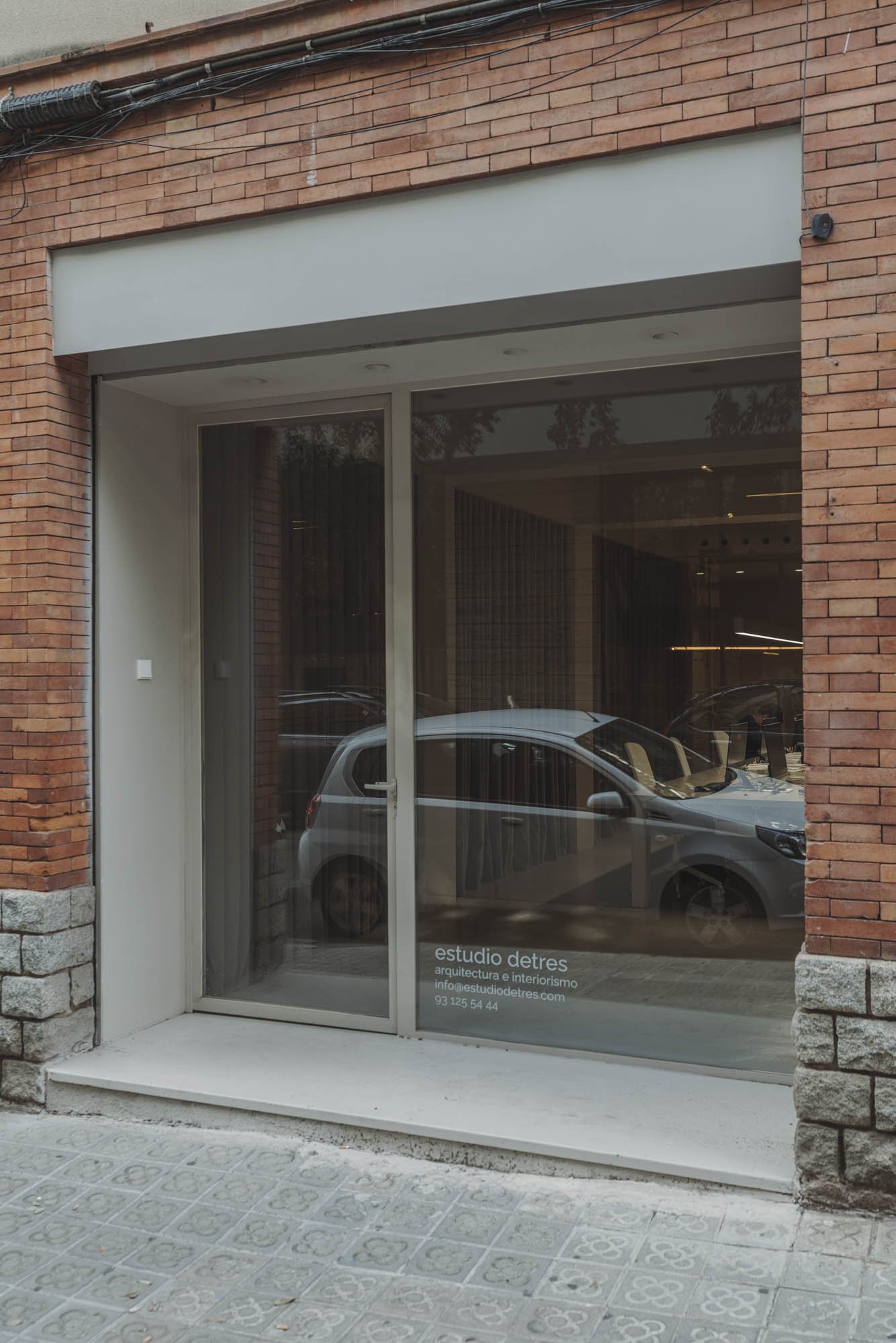 Fachada de un estudio de arquitectura con gran ventanal de vidrio y puertas corredizas, reflejando un coche y un árbol, con paredes de ladrillo y detalles en piedra en la base.