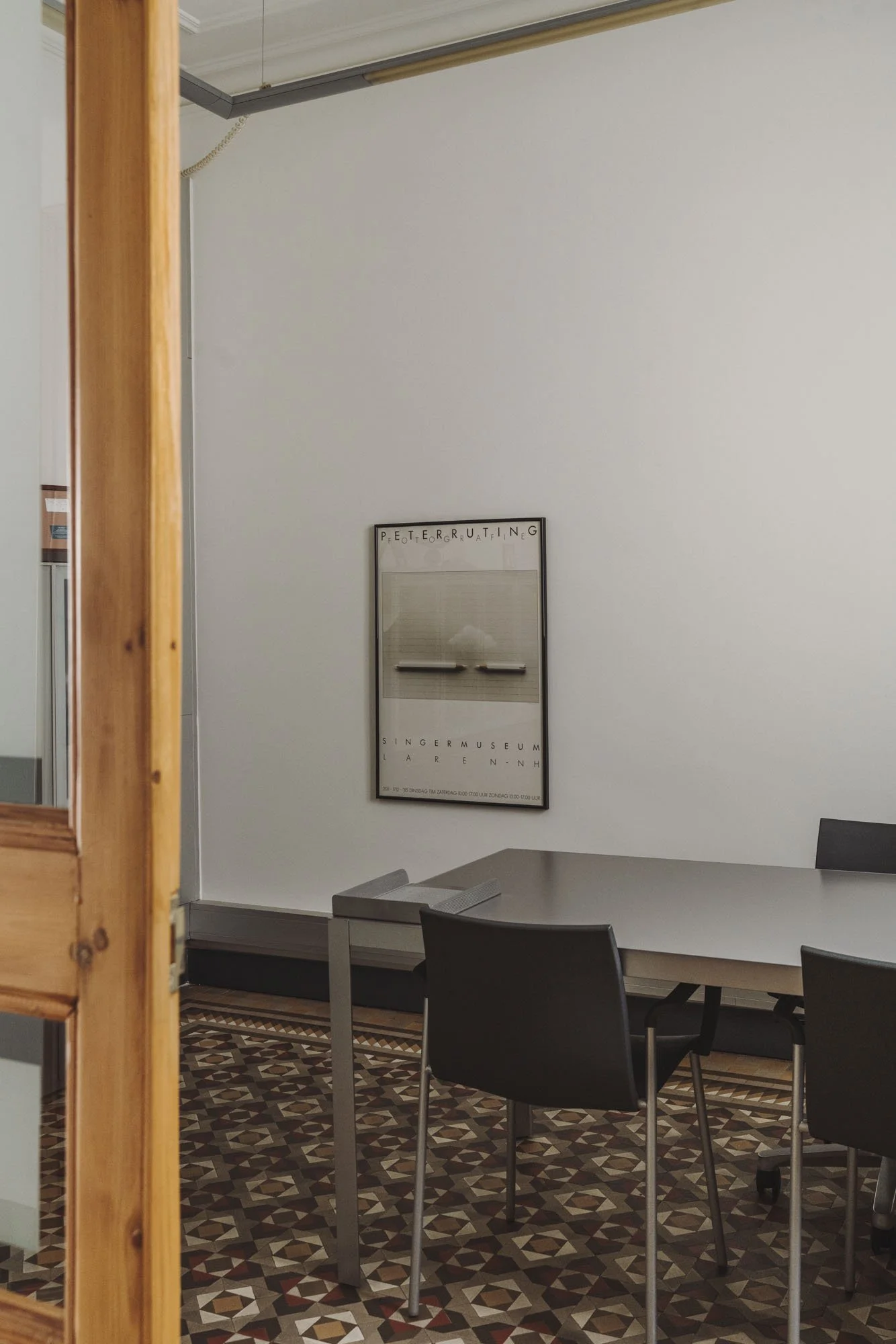 Vista de una sala con una mesa gris y varias sillas negras, en la pared hay un cartel con letras y un espejo, y en la esquina izquierda parte de una puerta de madera.