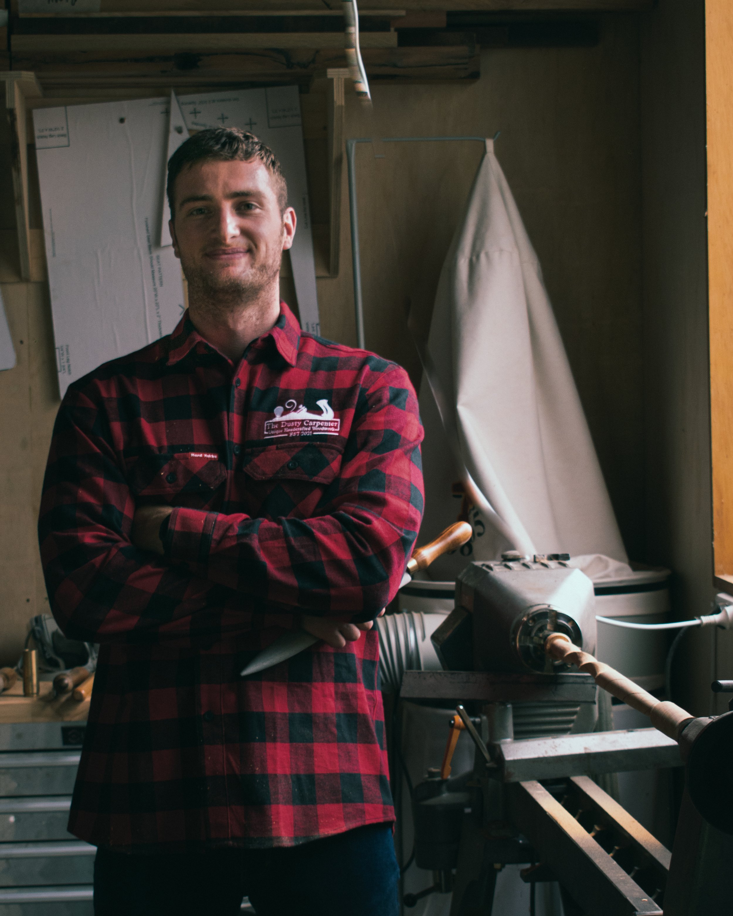 A man with short dark hair and a light beard wearing a red and black plaid shirt with a logo says "The Dusty Carpenter" on it, standing in a woodworking workshop. He is smiling and has his arms crossed. The background shows woodworking tools, a workbench, and a large machine for shaping wood.