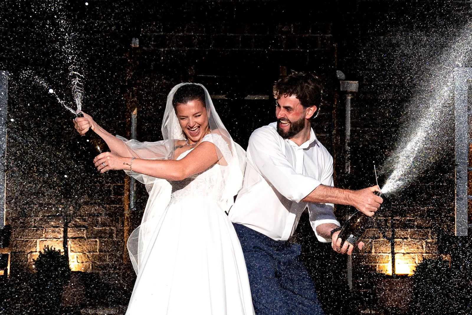 A bride and groom celebrating with champagne, spraying it in celebration, both smiling and enjoying the moment at their wedding reception.