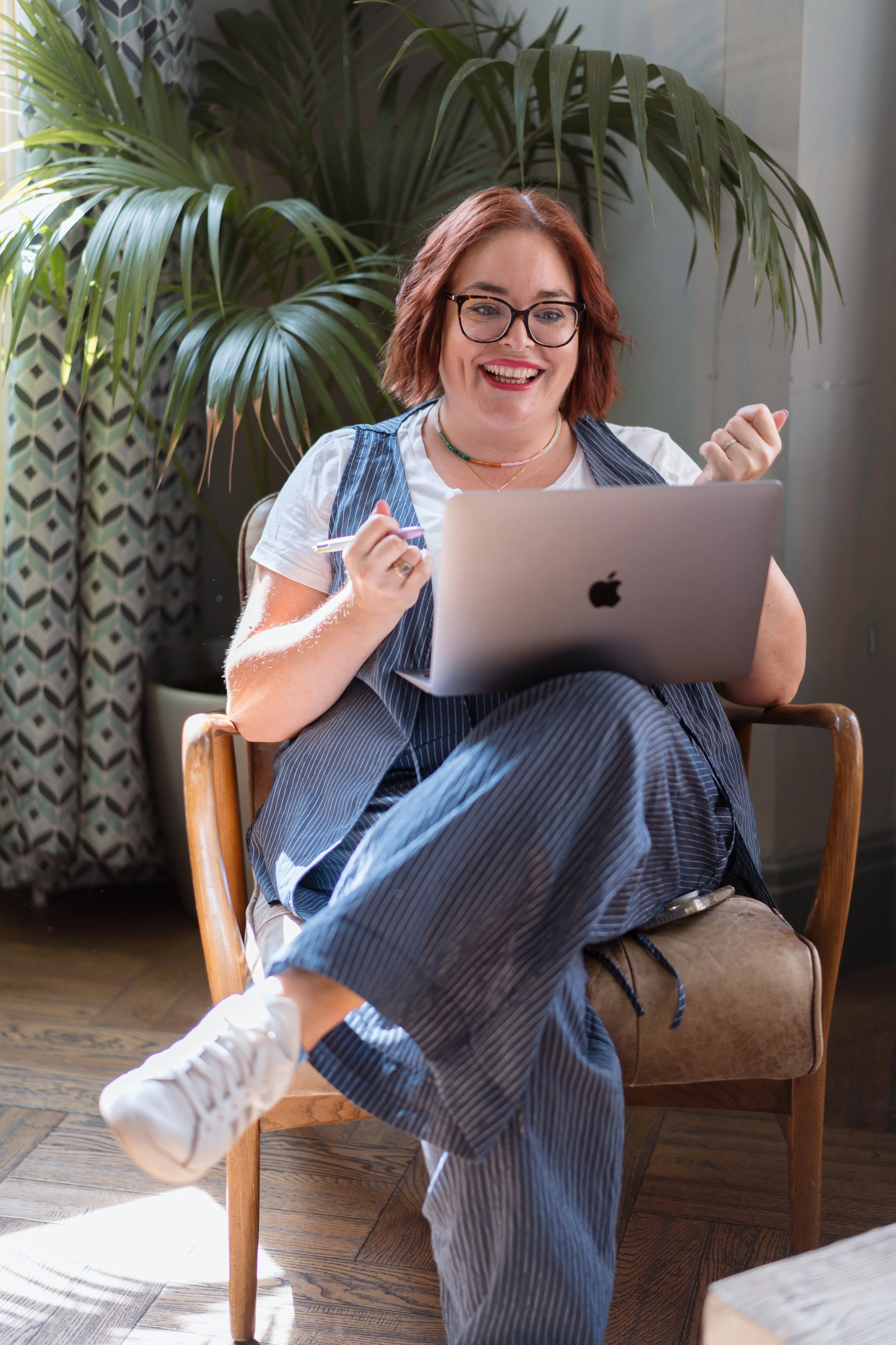 Jules Brim smiling excitedly while sitting in a wooden chair, holding a pen in one hand and working on her laptop, inside a room with large green plants and patterned curtains.