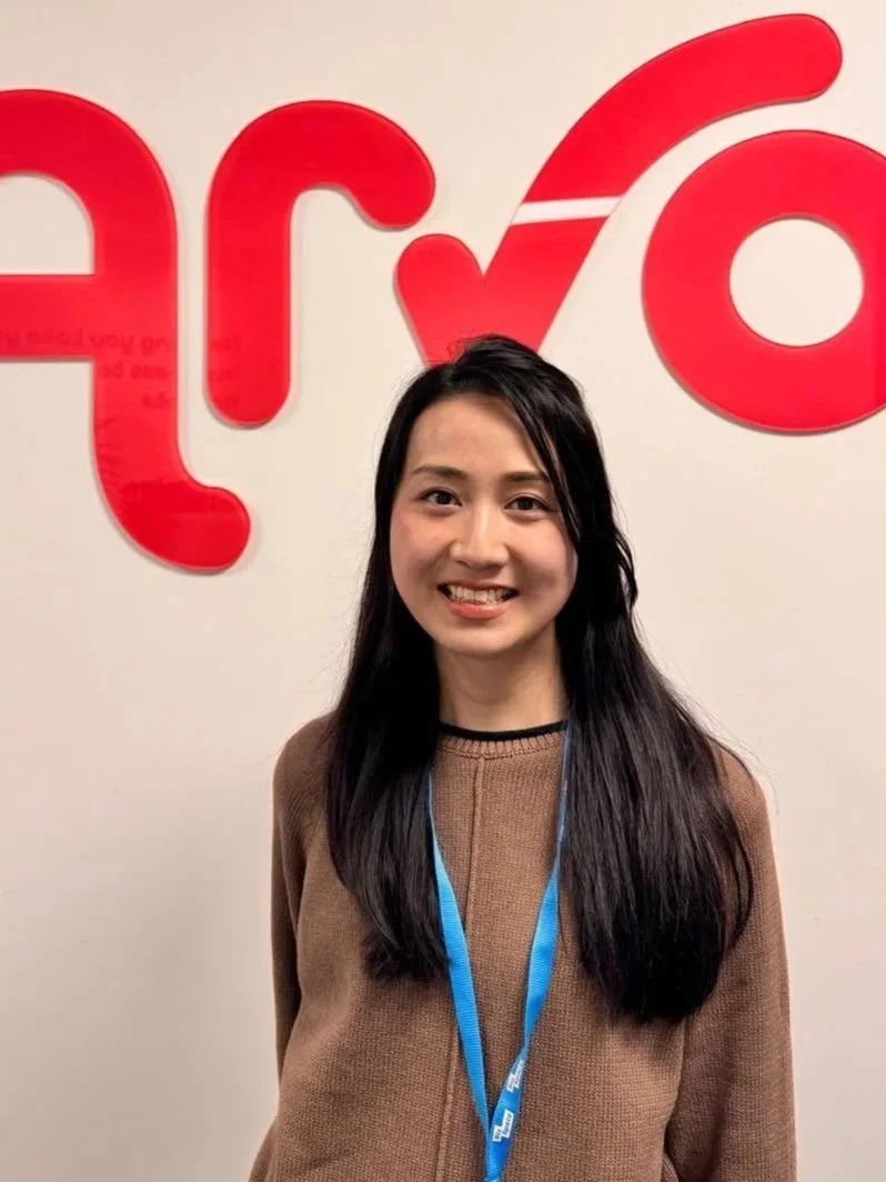 A young woman with long dark hair smiling in front of a beige wall with large red letters and a white stripe. She is wearing a brown sweater and a blue lanyard.