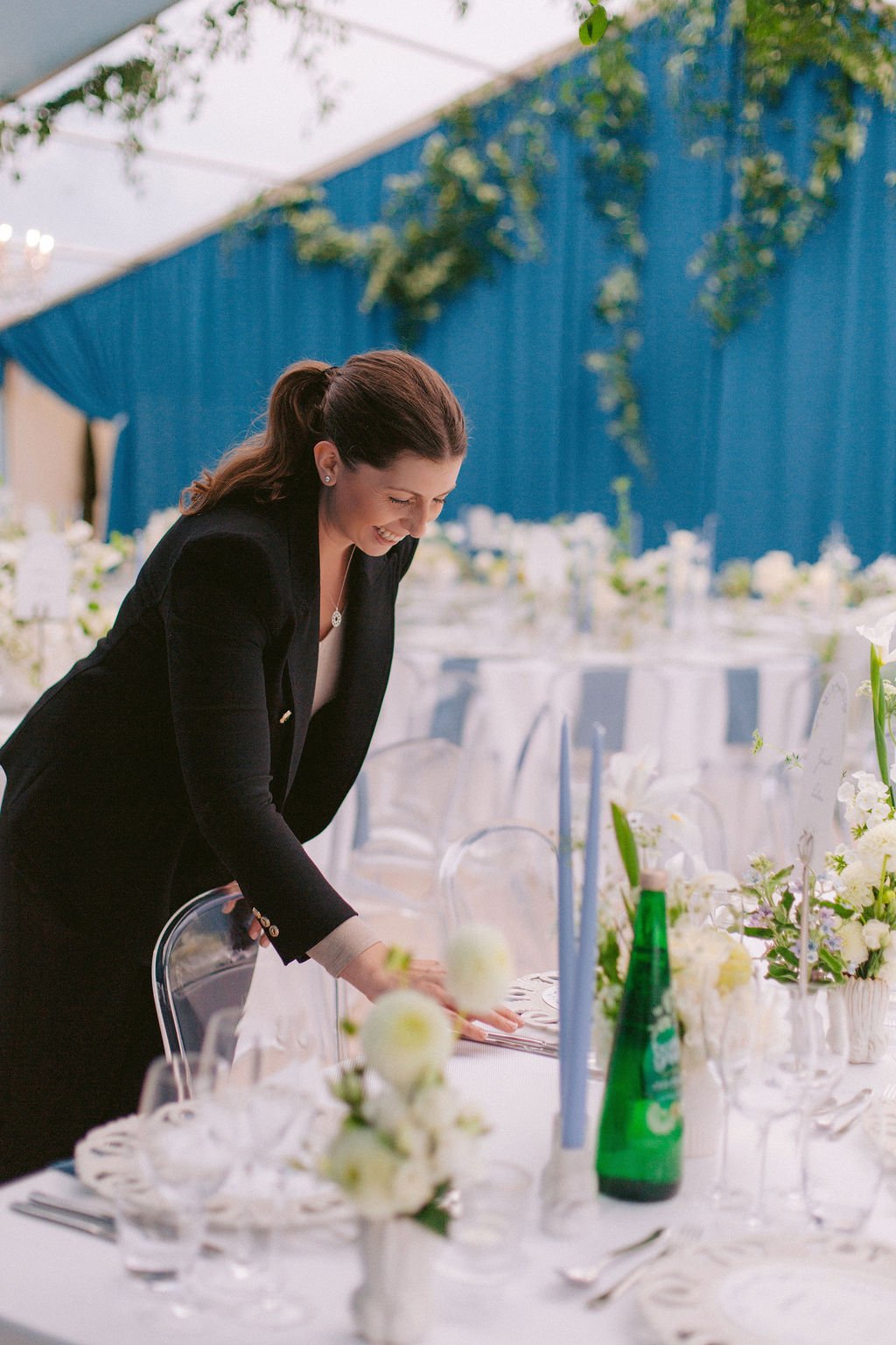 A woman in a black blazer setting a table with flowers and tableware at an event or celebration.