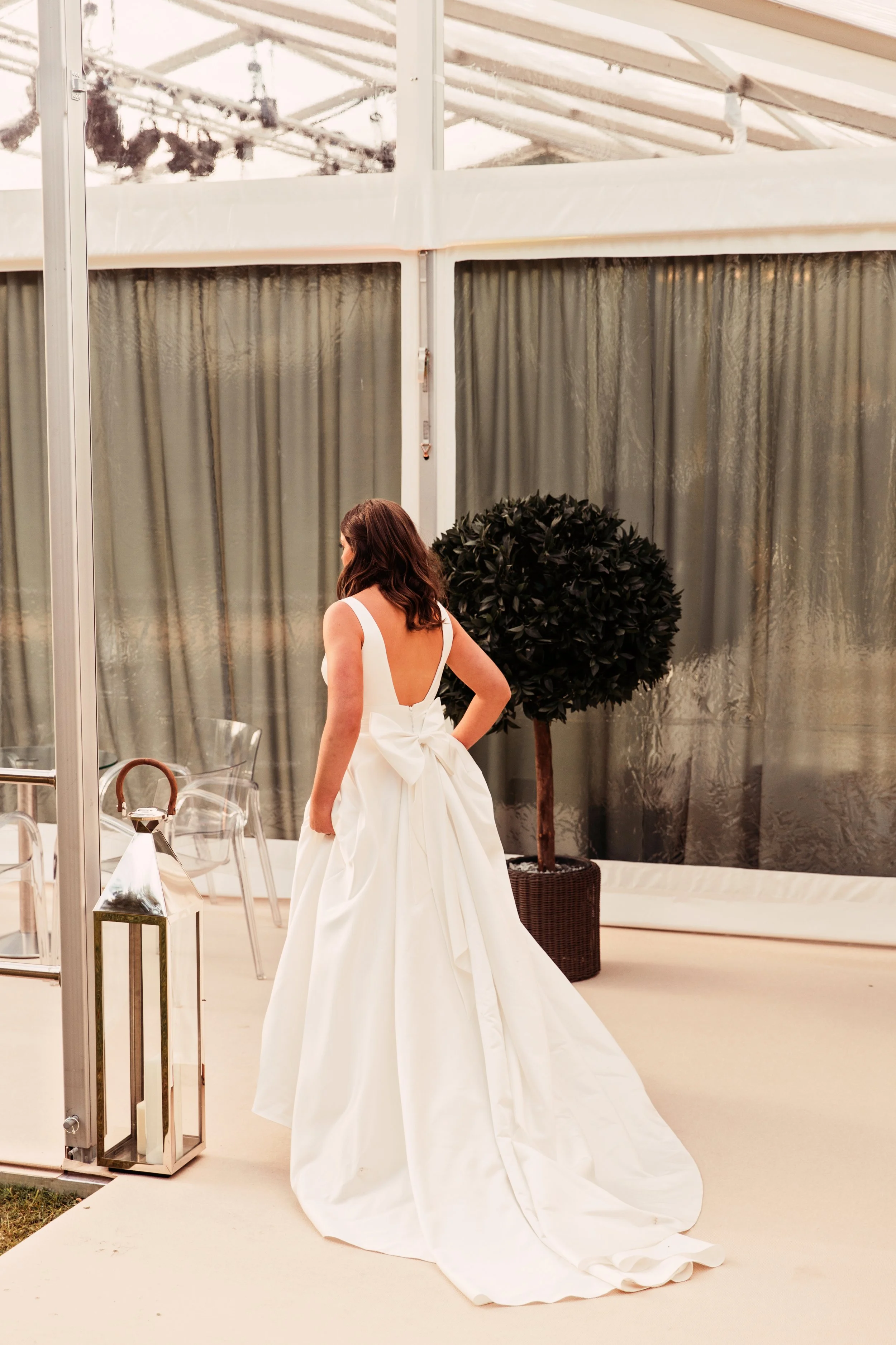 A woman in a white wedding dress with a large bow on the back, standing inside a glass-enclosed venue near a potted tree and clear chairs.