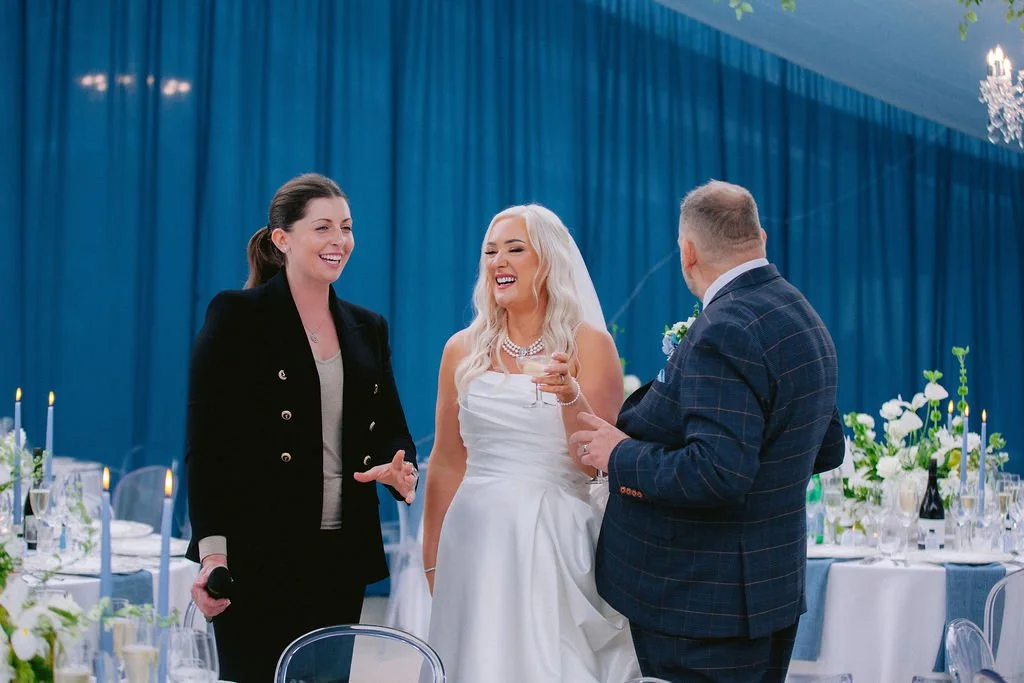 A bride with blond hair in a white wedding dress laughing and holding a glass of champagne, flanked by a woman in a black blazer and a man in a dark blue checkered suit, at a wedding reception with blue curtains and floral decorations.