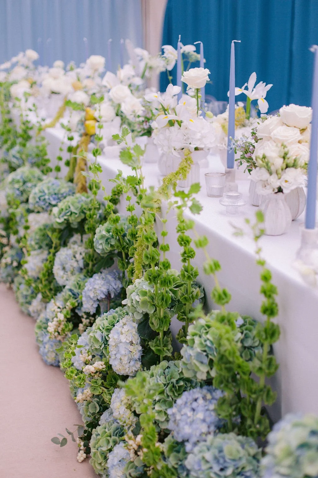 Table decorated with white flowers, including roses, lilies, and hydrangeas, with tall blue candles and glassware in a pastel-colored setting, likely for a wedding or special event.