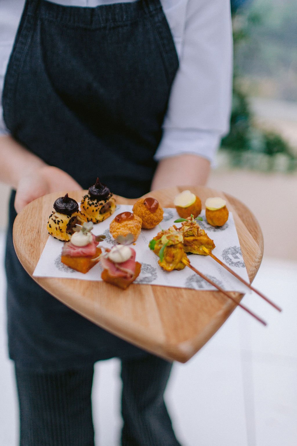 Person holding a wooden tray with various small, gourmet appetizers and desserts.