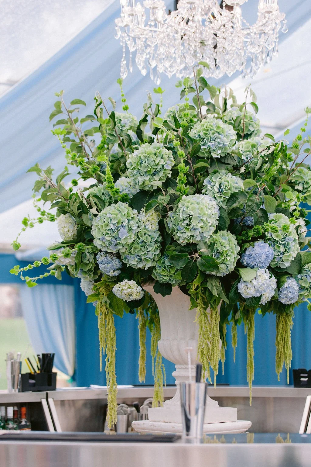 A large bouquet of light blue and white hydrangeas with green leaves and hanging greenery in a white decorative vase, positioned on a bar counter with glasses and bar tools visible in the foreground, under a crystal chandelier and blue drapes.