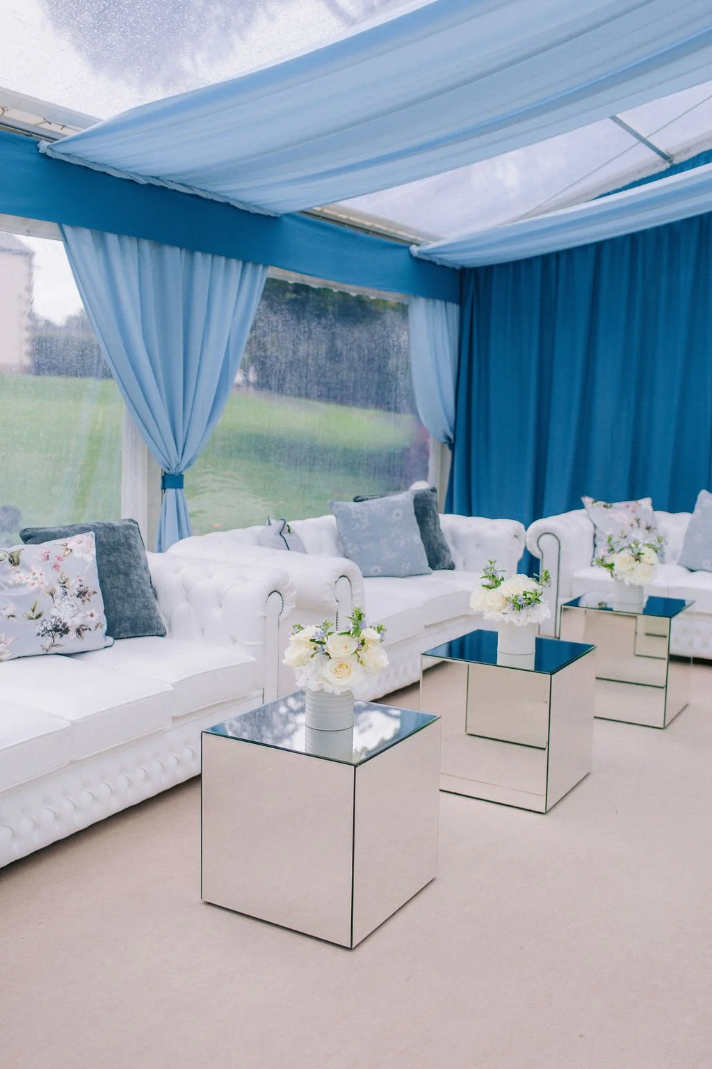 Interior of a stylish lounge area with white tufted sofas, floral and velvet cushions, mirrored side tables with white floral arrangements, and blue curtains and ceiling drapes, with a view of a green outdoor landscape through a rain-spattered window