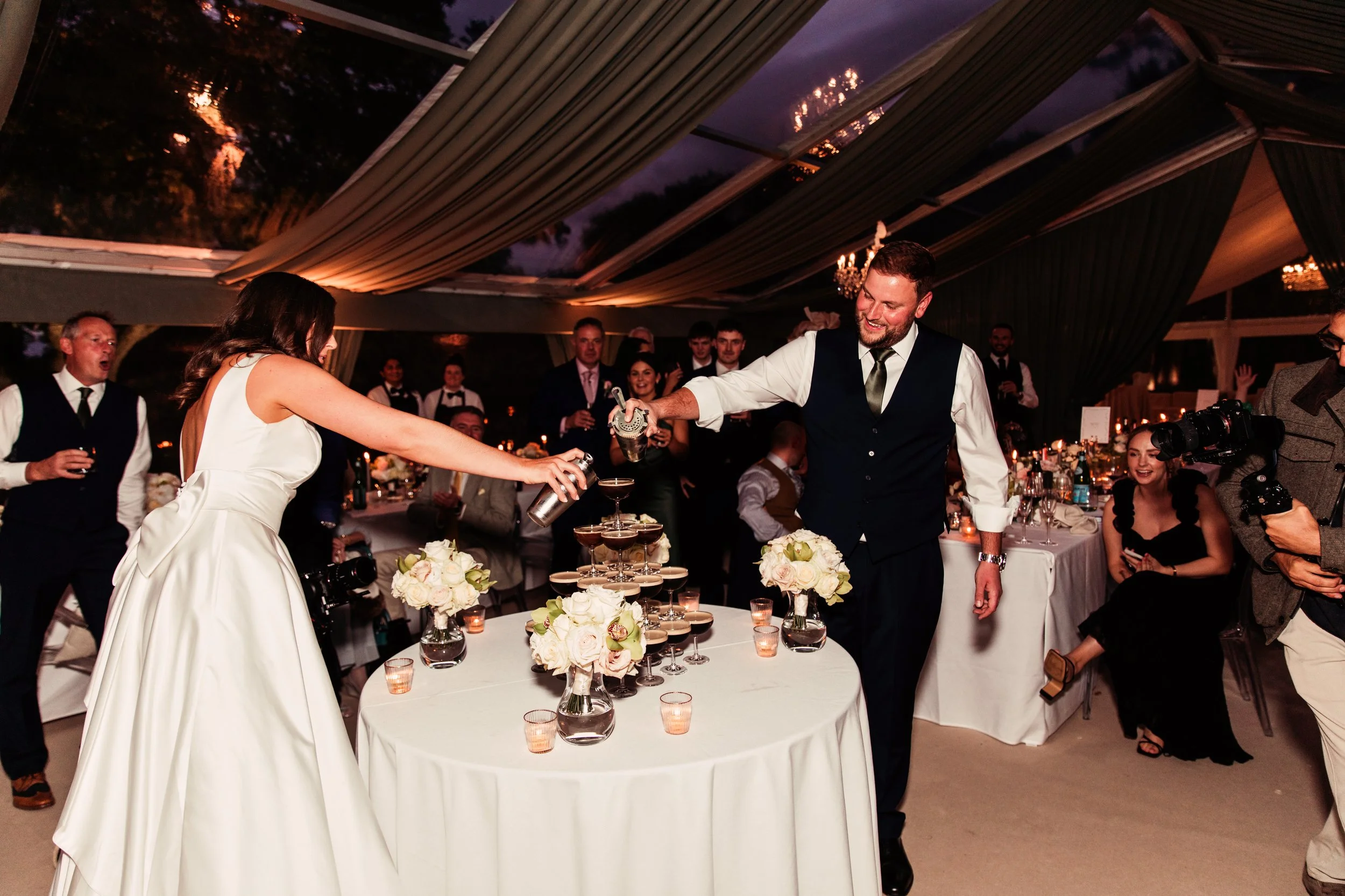 A bride and groom celebrating at their wedding reception, pouring champagne into a tower of glasses on a decorated table with white flowers, surrounded by guests in formal attire under a tent with draped fabric and chandeliers.
