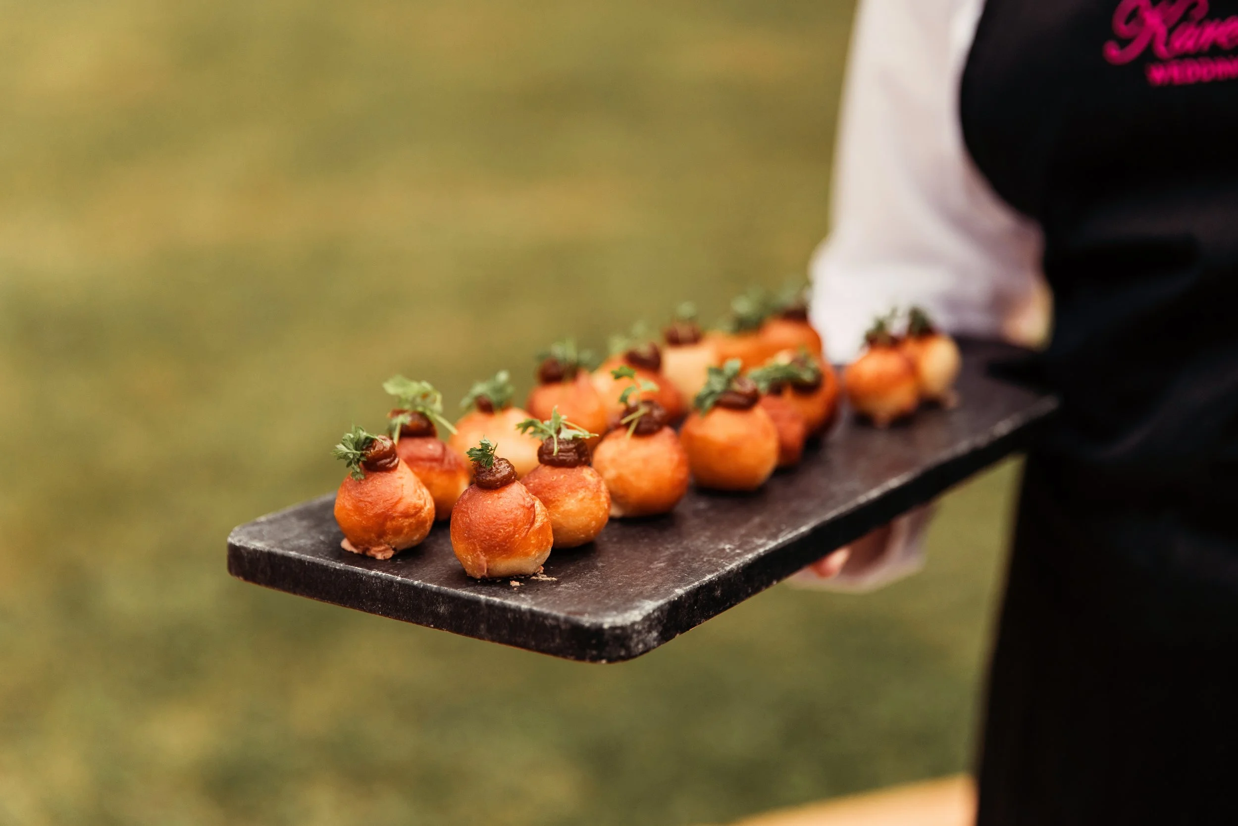 Person holding a black rectangular platter with small, round appetizers topped with herbs and a dollop of sauce, outdoors on a green grassy background.