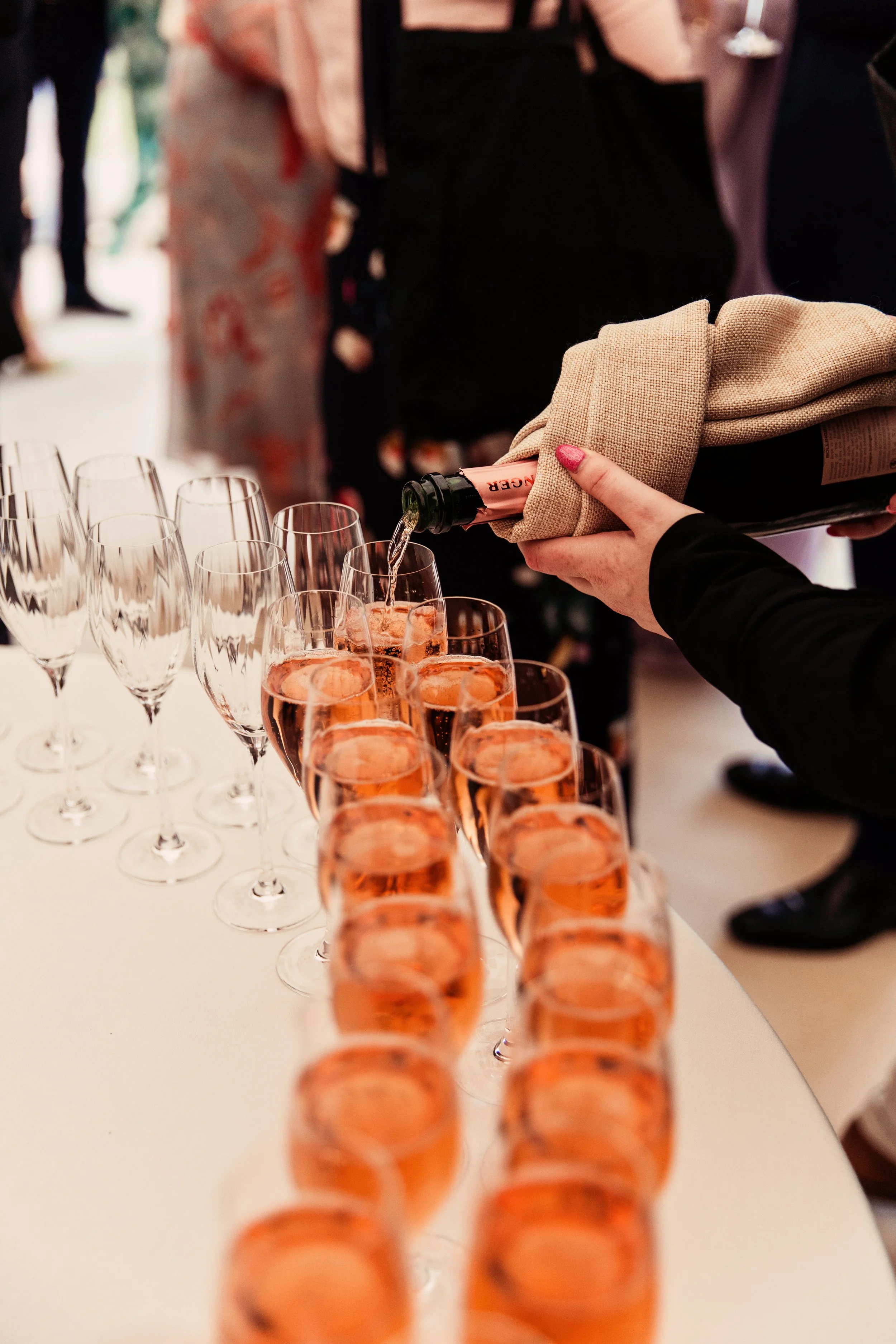 A person pouring rosé champagne from a bottle wrapped in a cloth into a row of filled champagne flutes at a celebration or event.
