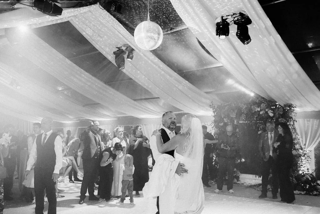 A bride and groom share their first dance at a wedding reception with guests in the background, decorated with draped fabric and a disco ball.