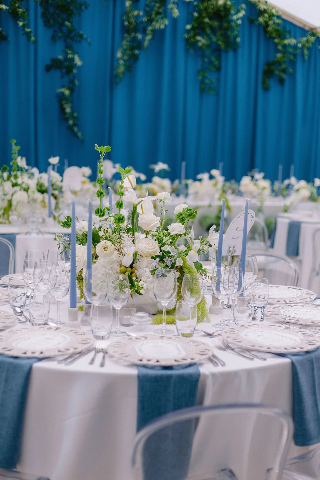 Formal event setup with a round table decorated with a white and blue color scheme, featuring a large floral centerpiece of white flowers and greenery, candles, and place settings with plates, glassware, and cutlery. Blue curtains with green vines in
