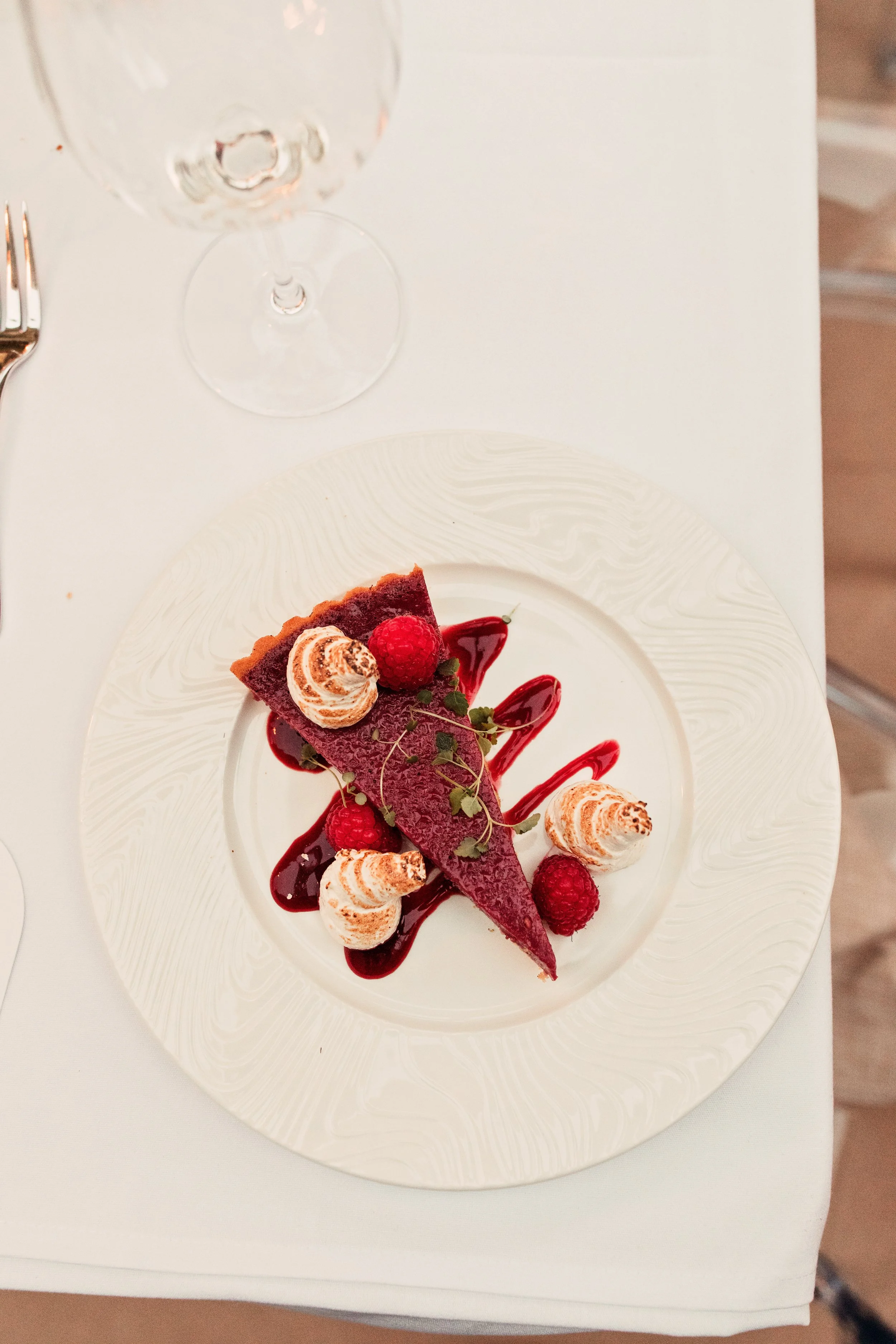 A slice of berry tart with raspberries, mint leaves, whipped cream, and berry sauce on a white decorative plate.