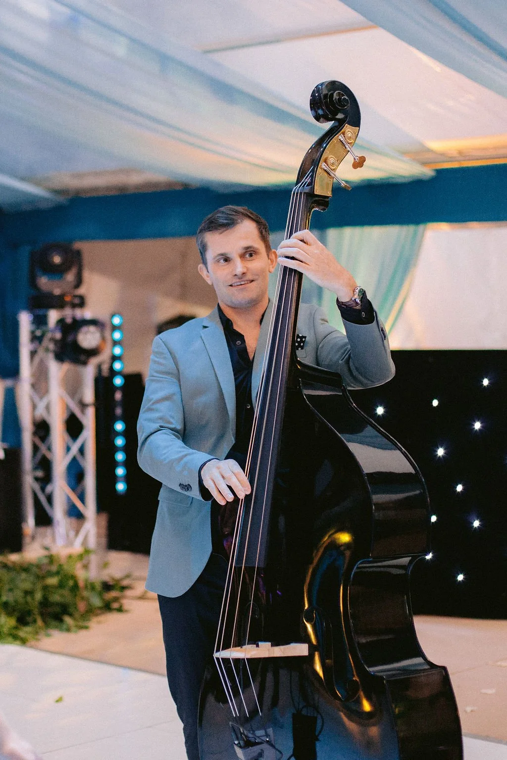 A man in a grey blazer playing a double bass at an indoor event with stage lighting and decorative elements in the background.