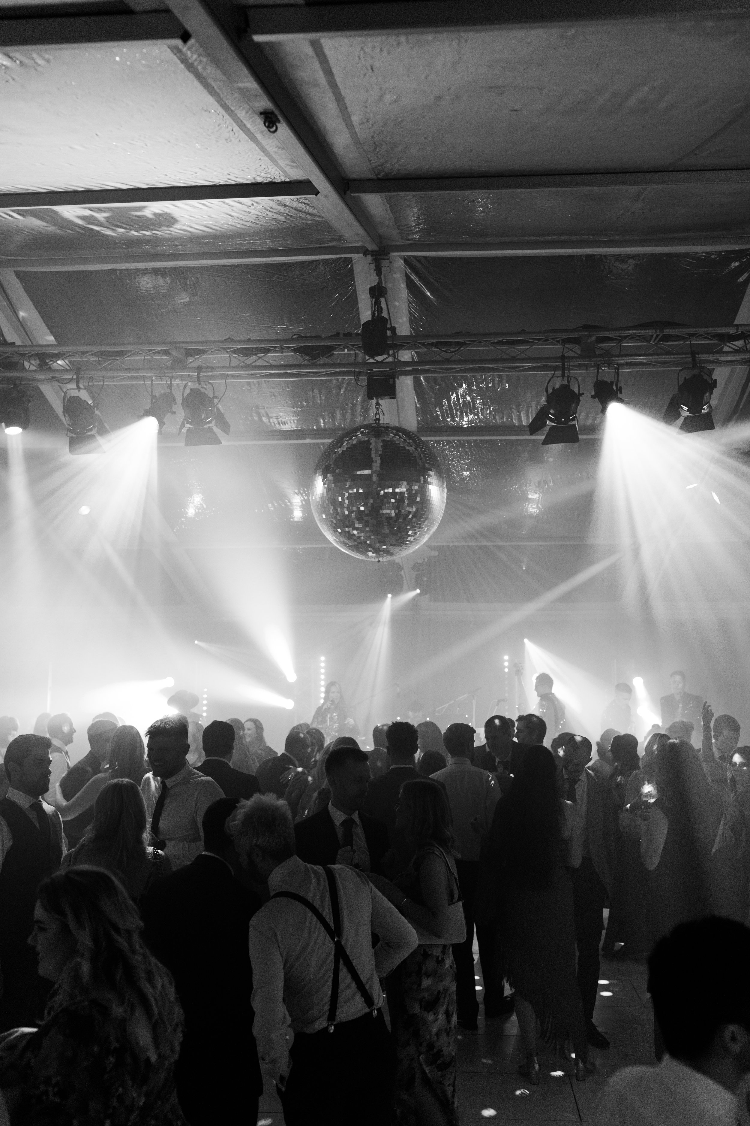Black and white photo of a crowded dance floor in a nightclub, with people dancing and socializing under a disco ball and stage lights.