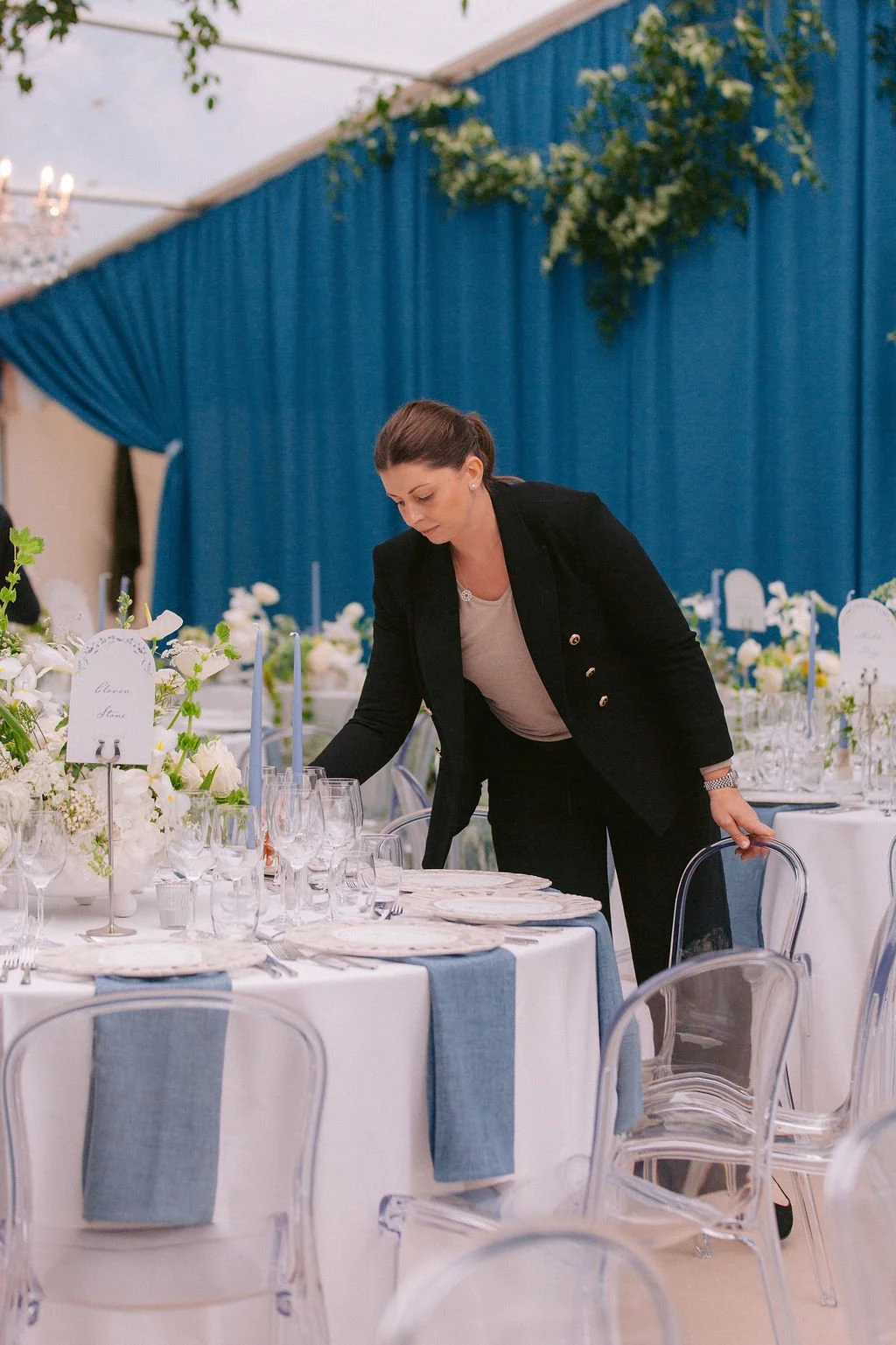 A woman in a black blazer and white shirt setting a formal dining table with white flowers, blue napkins, and glassware in a decorated event space with blue curtains.