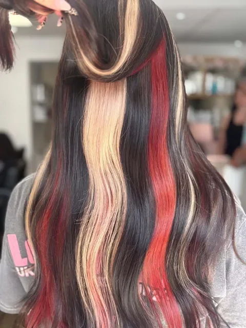 Back view of a woman with wavy, shoulder-length hair in a salon. A person with a tattoo on her arm is touching her hair.