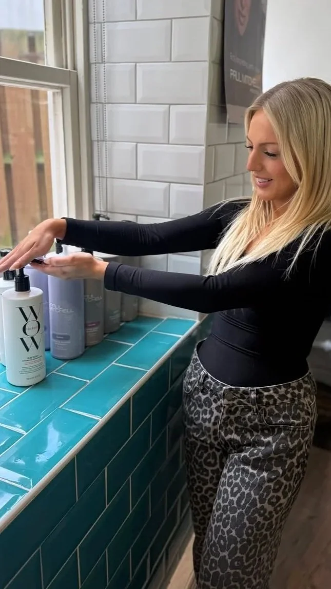 A woman with blonde hair is arranging or decorating a shelf with ivy vines and books in a room with a brick wall and window.
