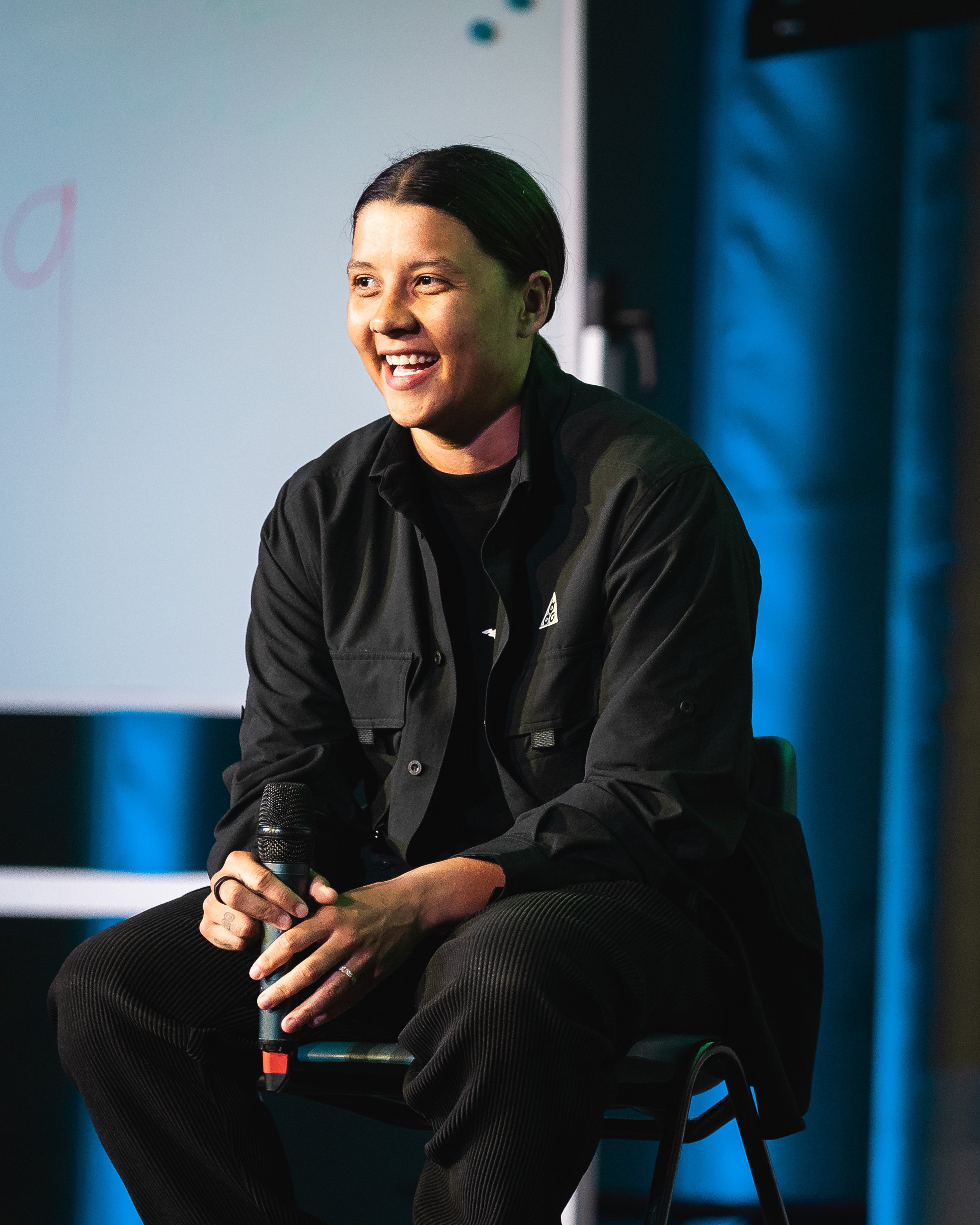A woman with dark hair sitting on a black chair, smiling, holding a microphone, wearing a black jacket, in a setting with a blue background and a white screen.