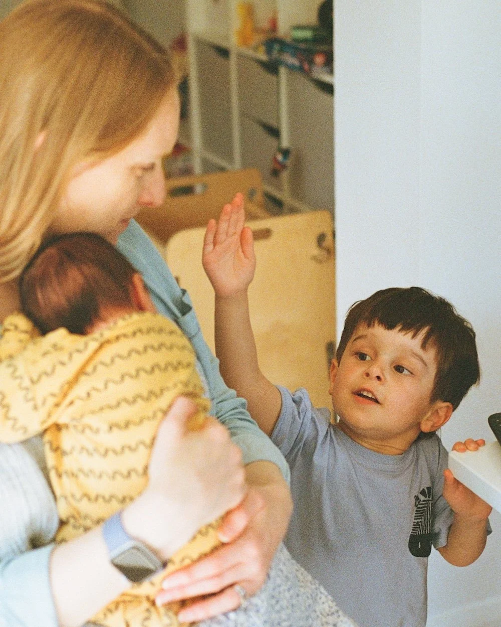 A young boy reaches up to his mother and baby brother as they sit at a kitchen chair above him by London photographer Bobby Shoebotham 