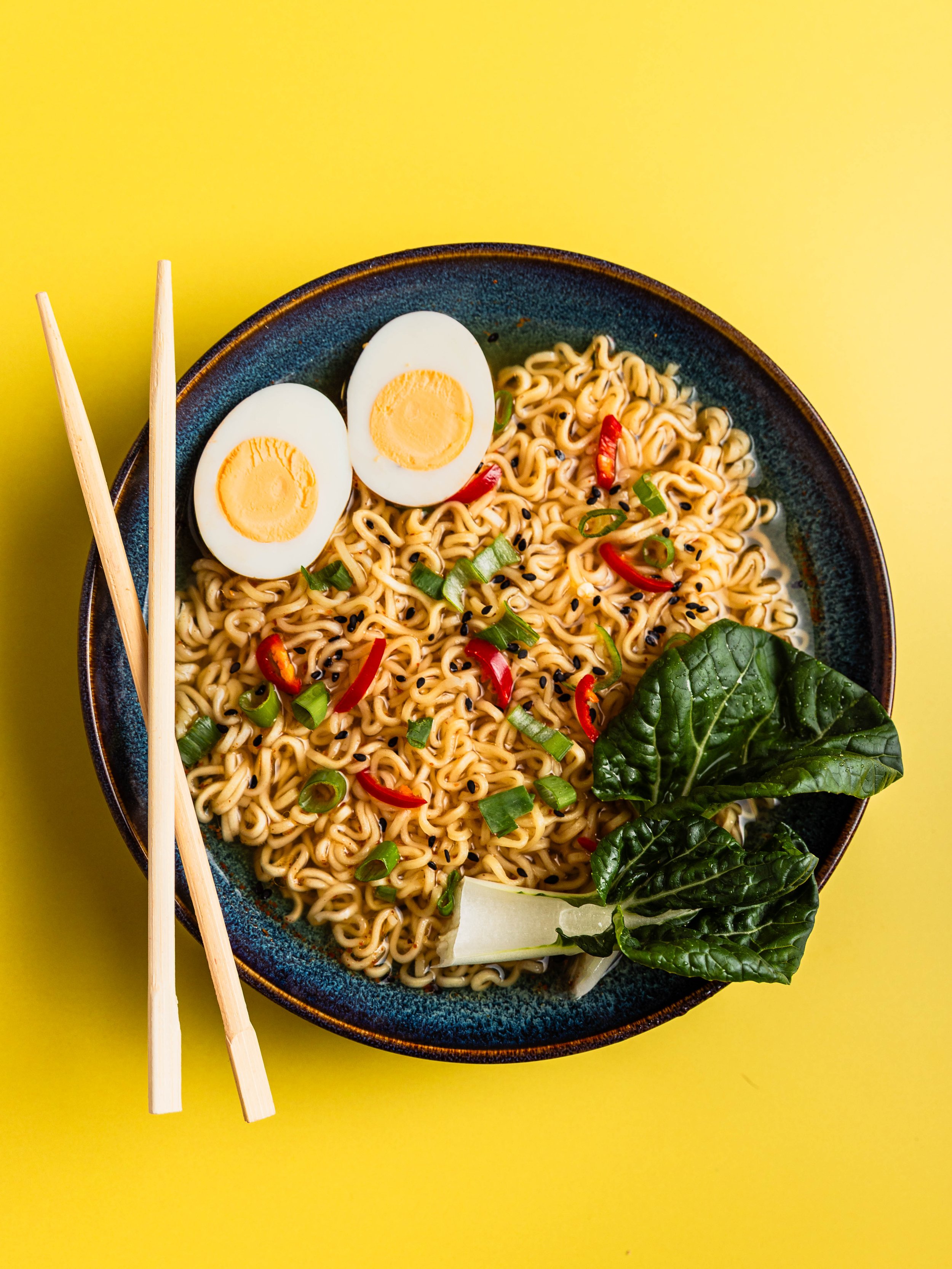 A bowl of ramen noodles with sliced boiled eggs, chopped green onions, red chili slices, and leafy greens on a yellow background with chopsticks resting on the side.