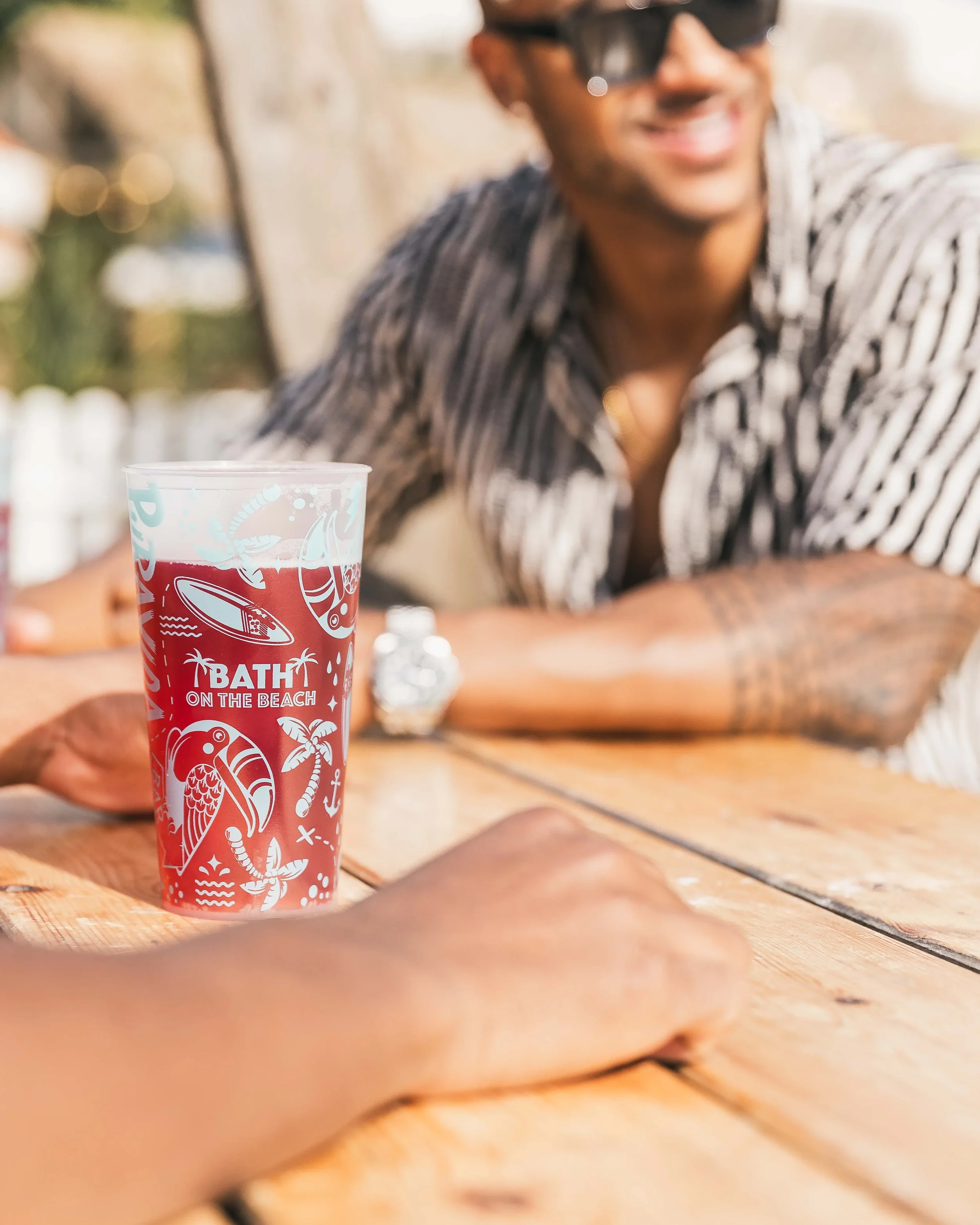 A colorful drink with a straw on a wooden table in front of a smiling man with tattoos, glasses, and a striped shirt, sitting outdoors.