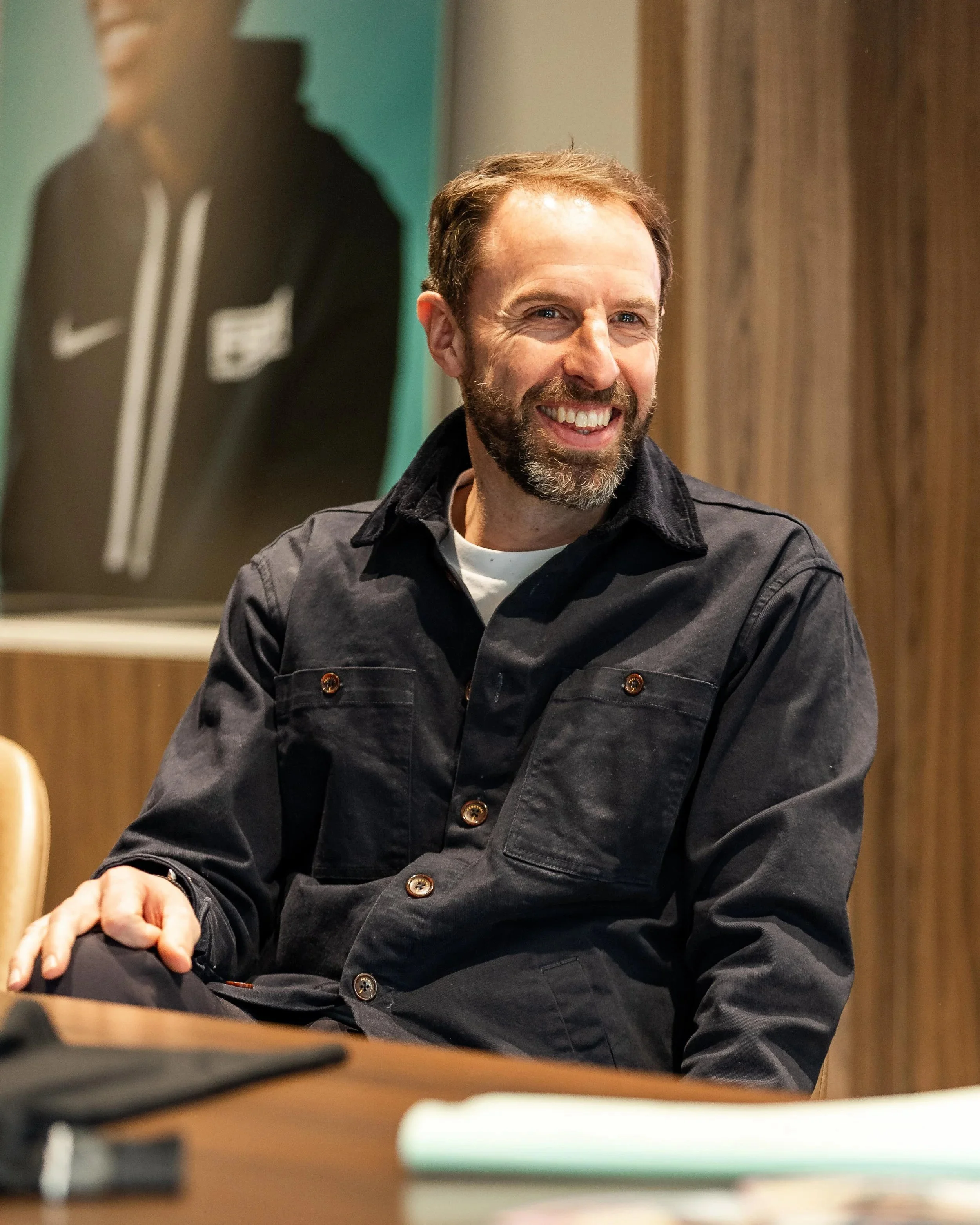 A smiling man with a beard and short brown hair, wearing a black jacket, sitting at a wooden table in a room with wood-paneled walls and a large poster in the background.