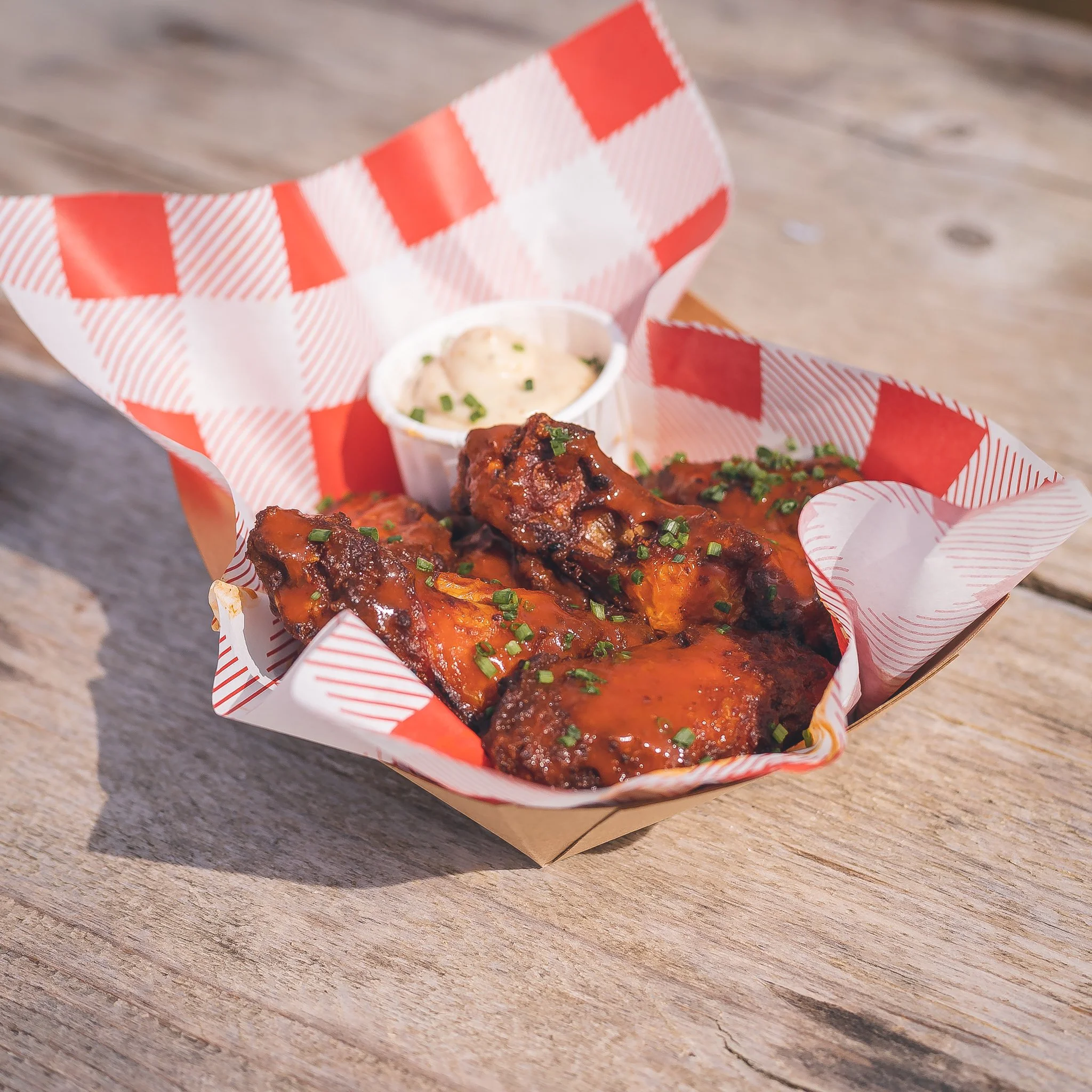 A paper food tray with buffalo chicken wings garnished with chopped green onions, served with a side of ranch dressing in a small cup, placed on a wooden surface.