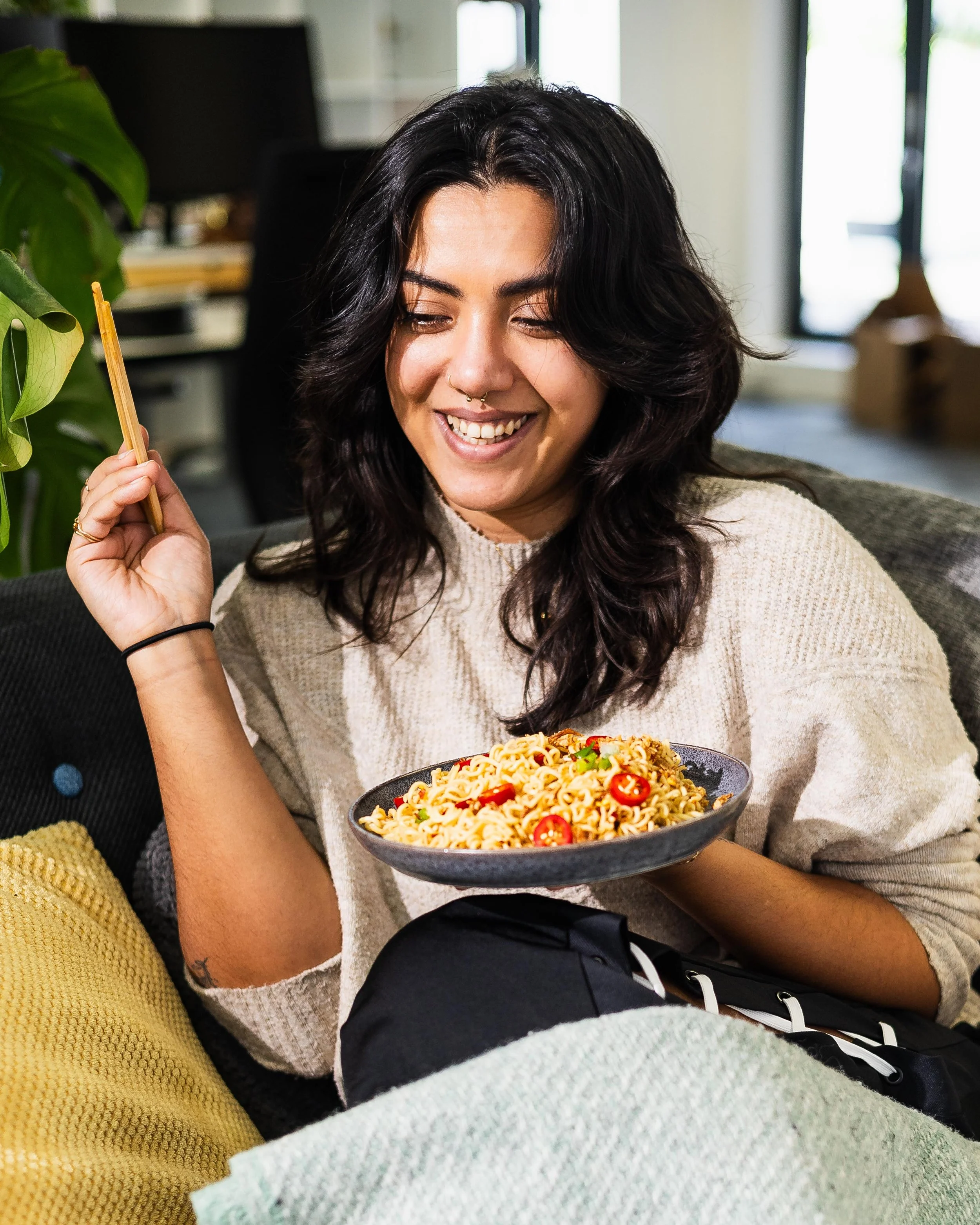 A woman with dark, wavy hair smiling and holding a plate of Asian noodles topped with sliced chili peppers, sitting on a couch in a cozy room.