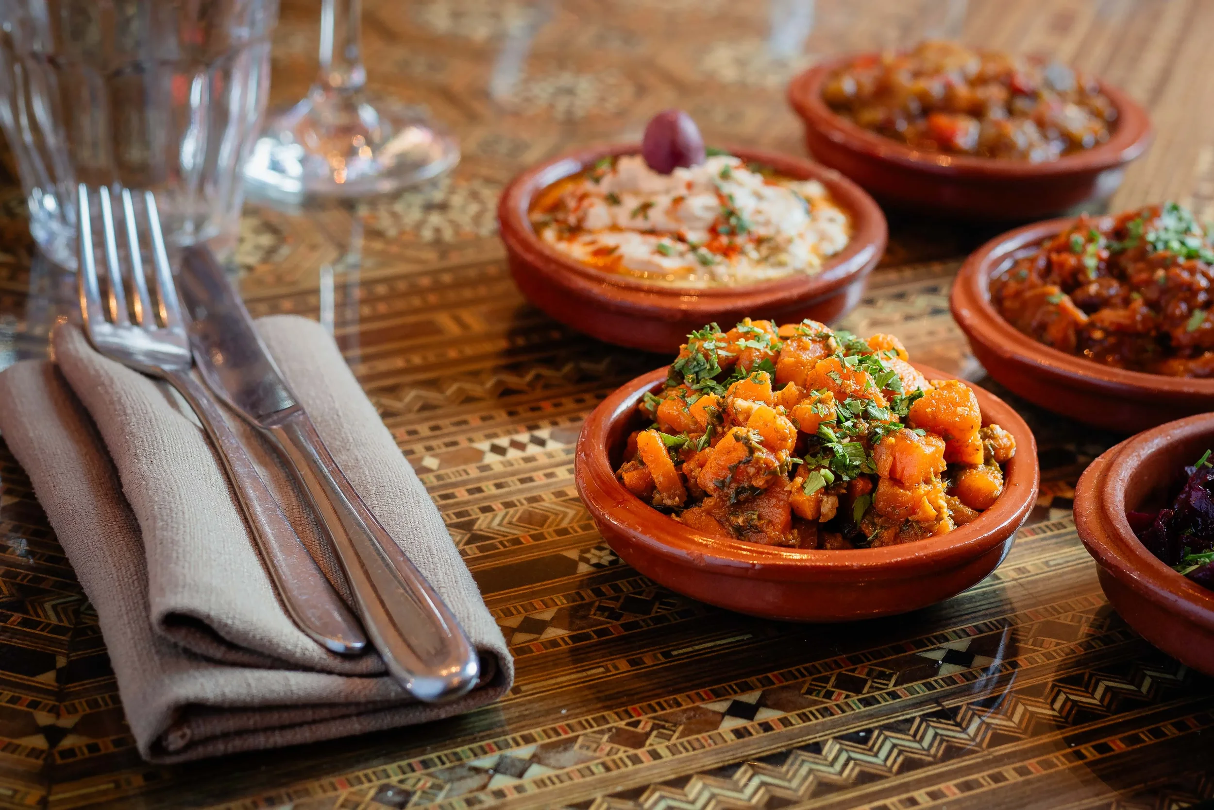 A table set with traditional Mexican food in small terracotta bowls, including a spicy vegetable dish and other flavorful stews, with a fork and knife on a beige napkin and glasses nearby.