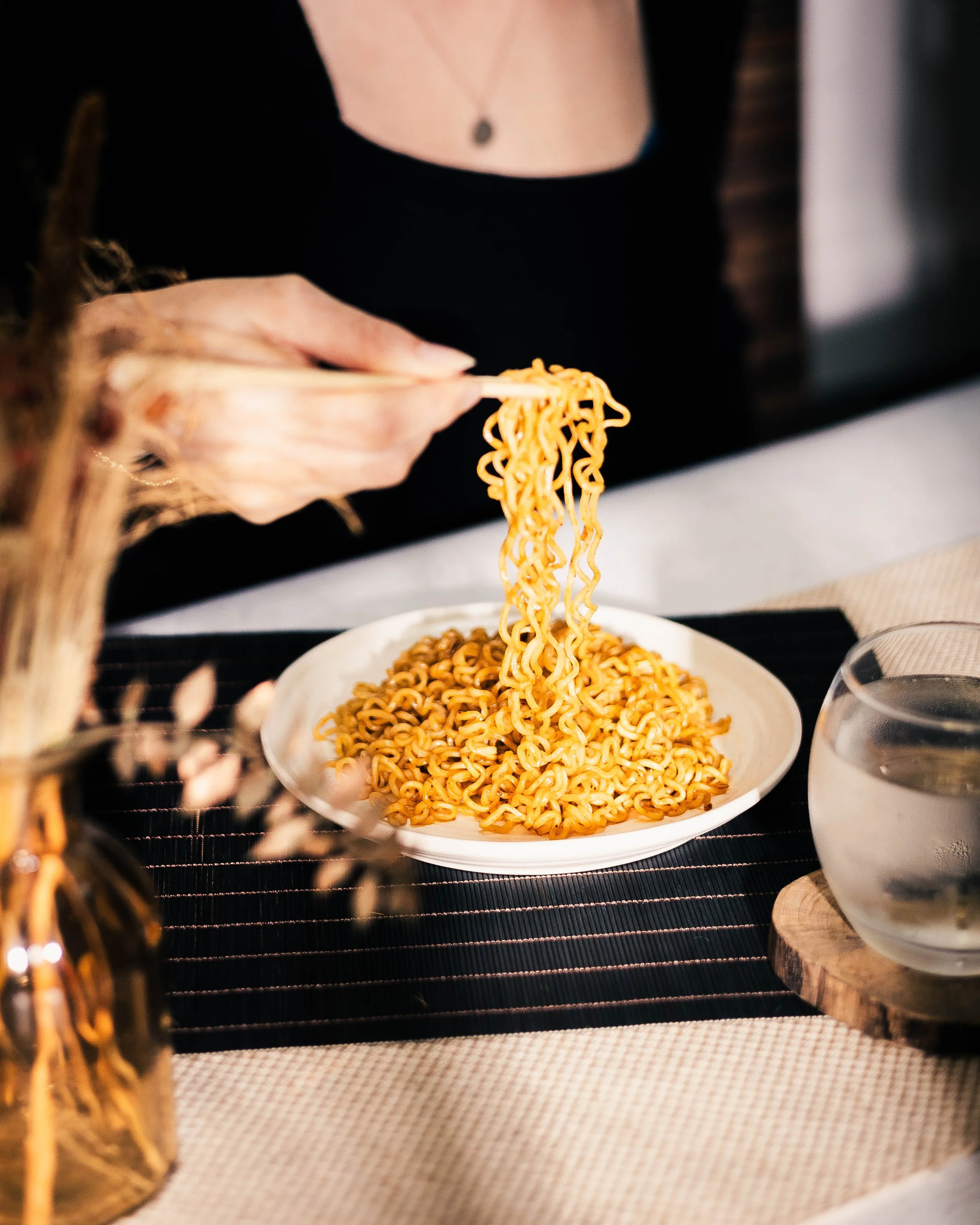Person lifting cooked ramen noodles with chopsticks from a white plate on a black placemat, with a glass of water on the side.