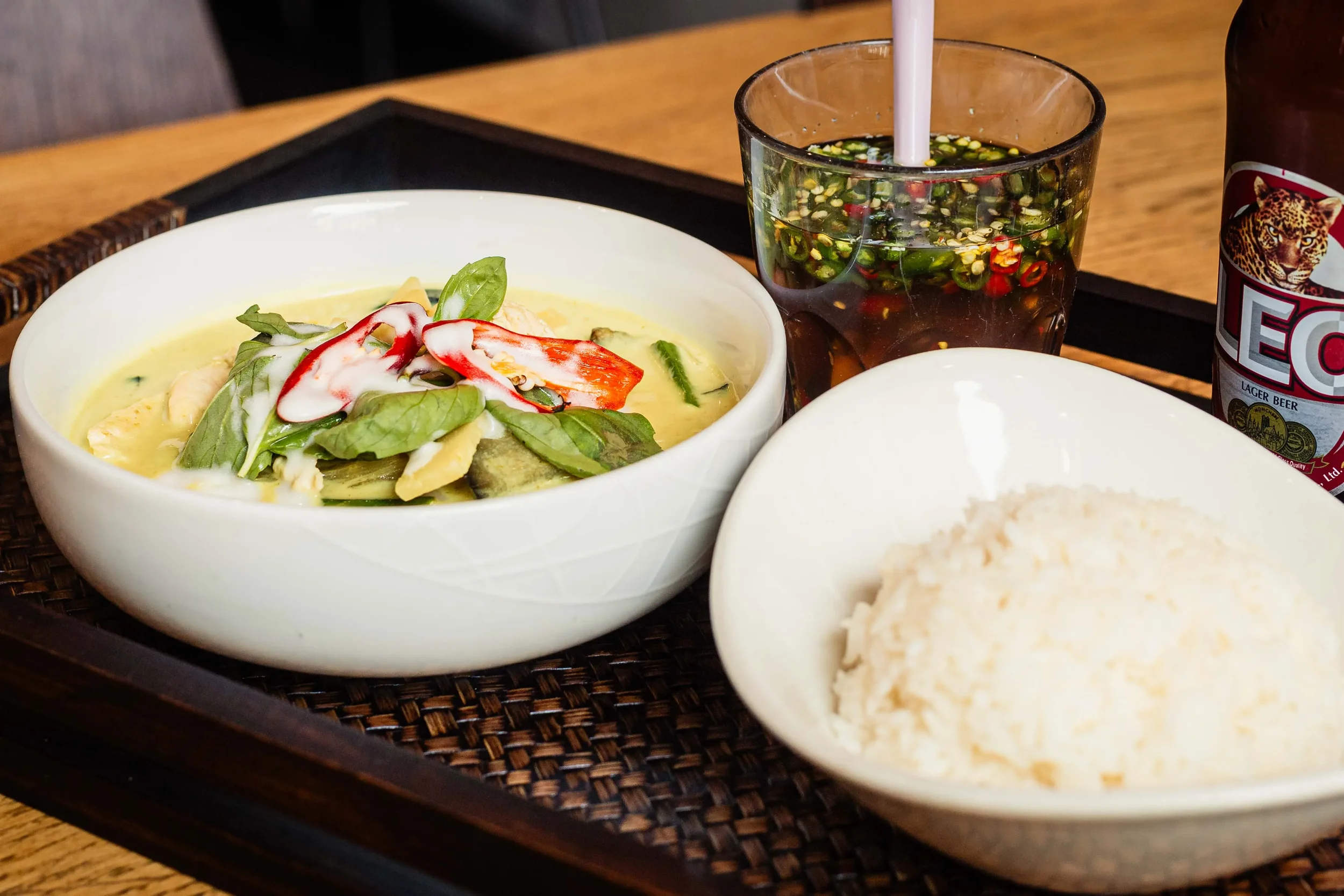 A tray with a bowl of curry topped with herbs and chili slices, a bowl of white rice, a glass of soy sauce with chopped chili, and a bottle of beer.