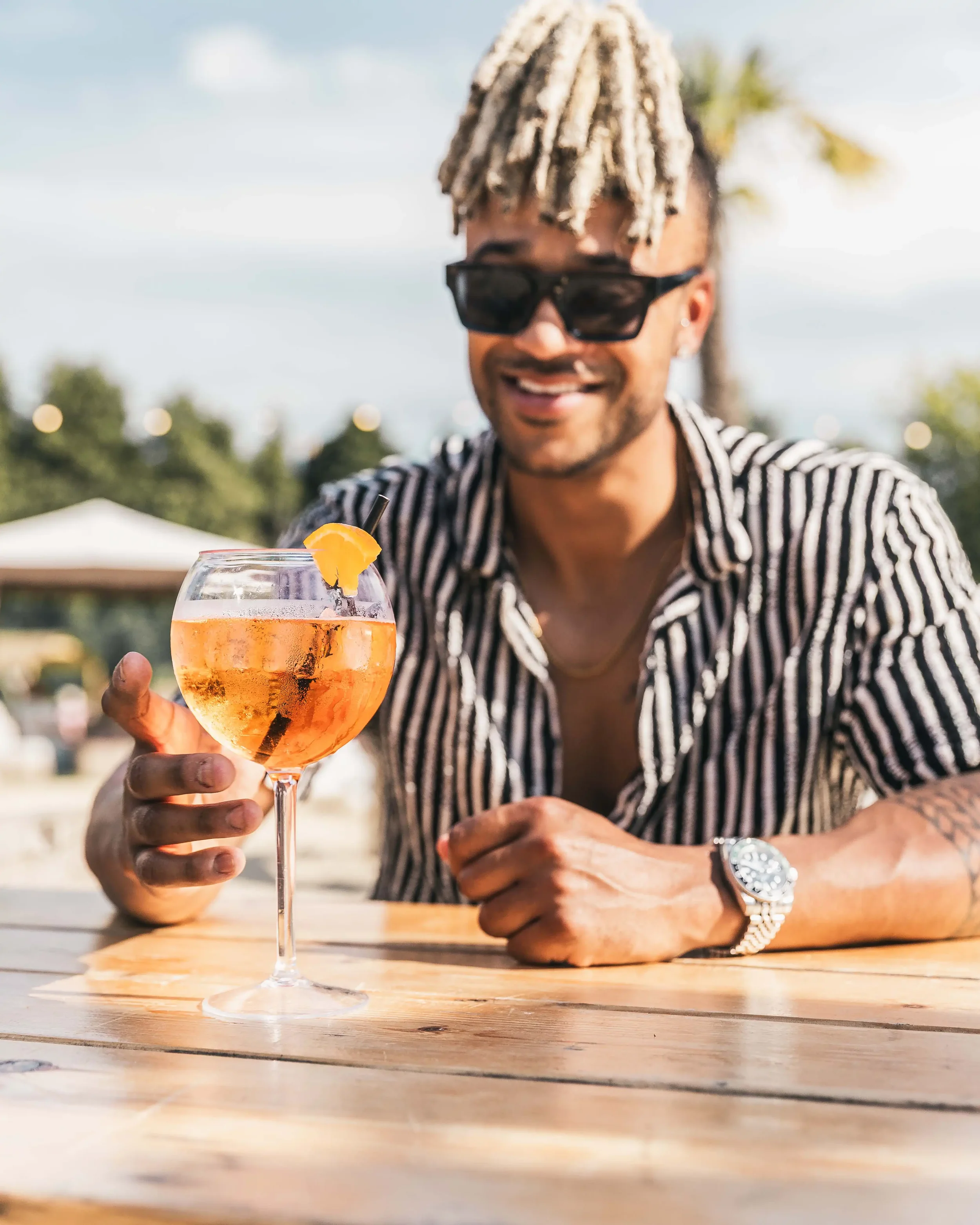 A man with sunglasses and striped shirt sitting at an outdoor table, holding a cocktail glass with orange-colored drink, a lemon slice garnish, and a black straw, smiling at the camera.