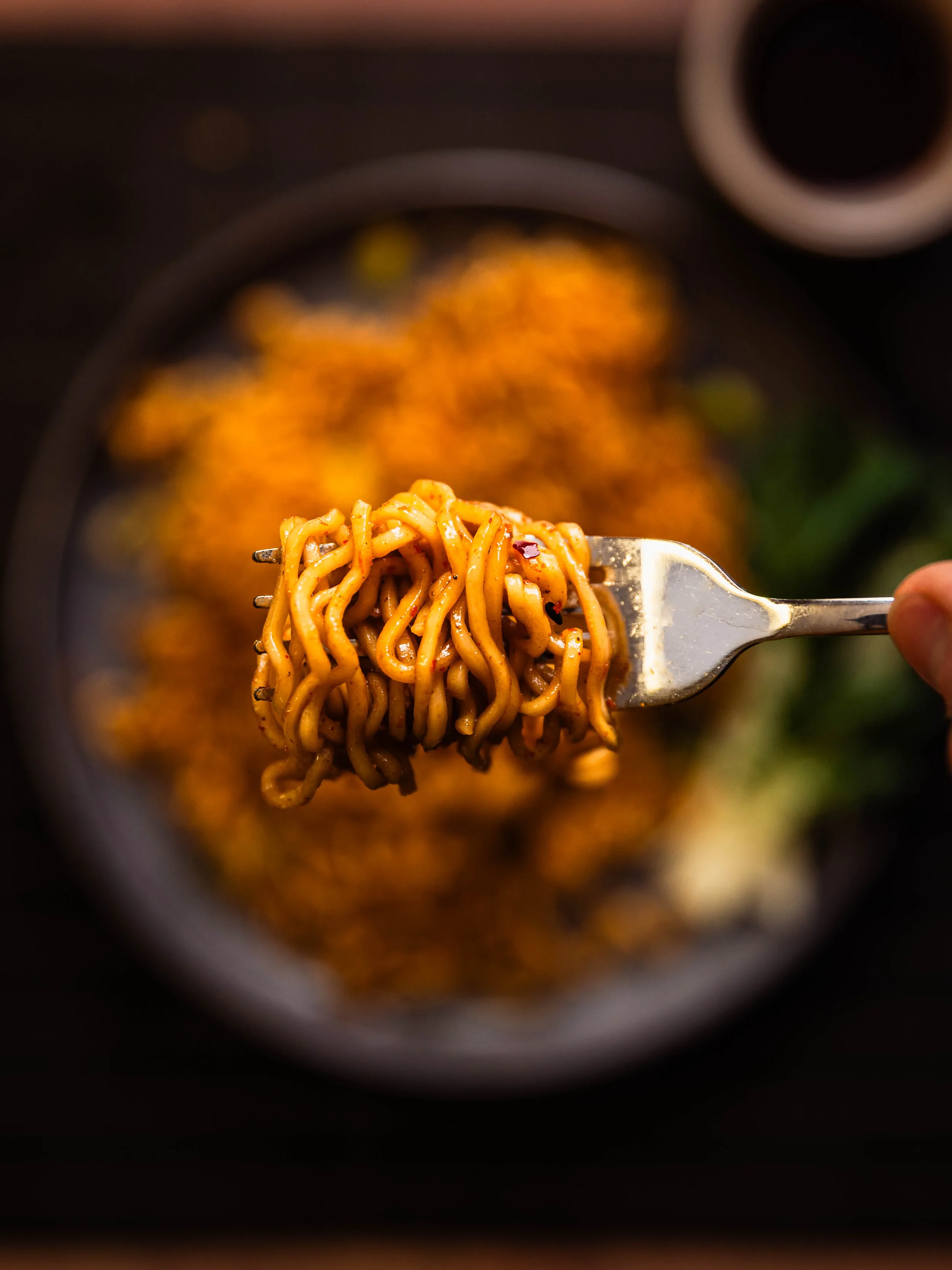 Close-up of a fork lifting cooked ramen noodles with a blurred background of more noodles and a bowl.