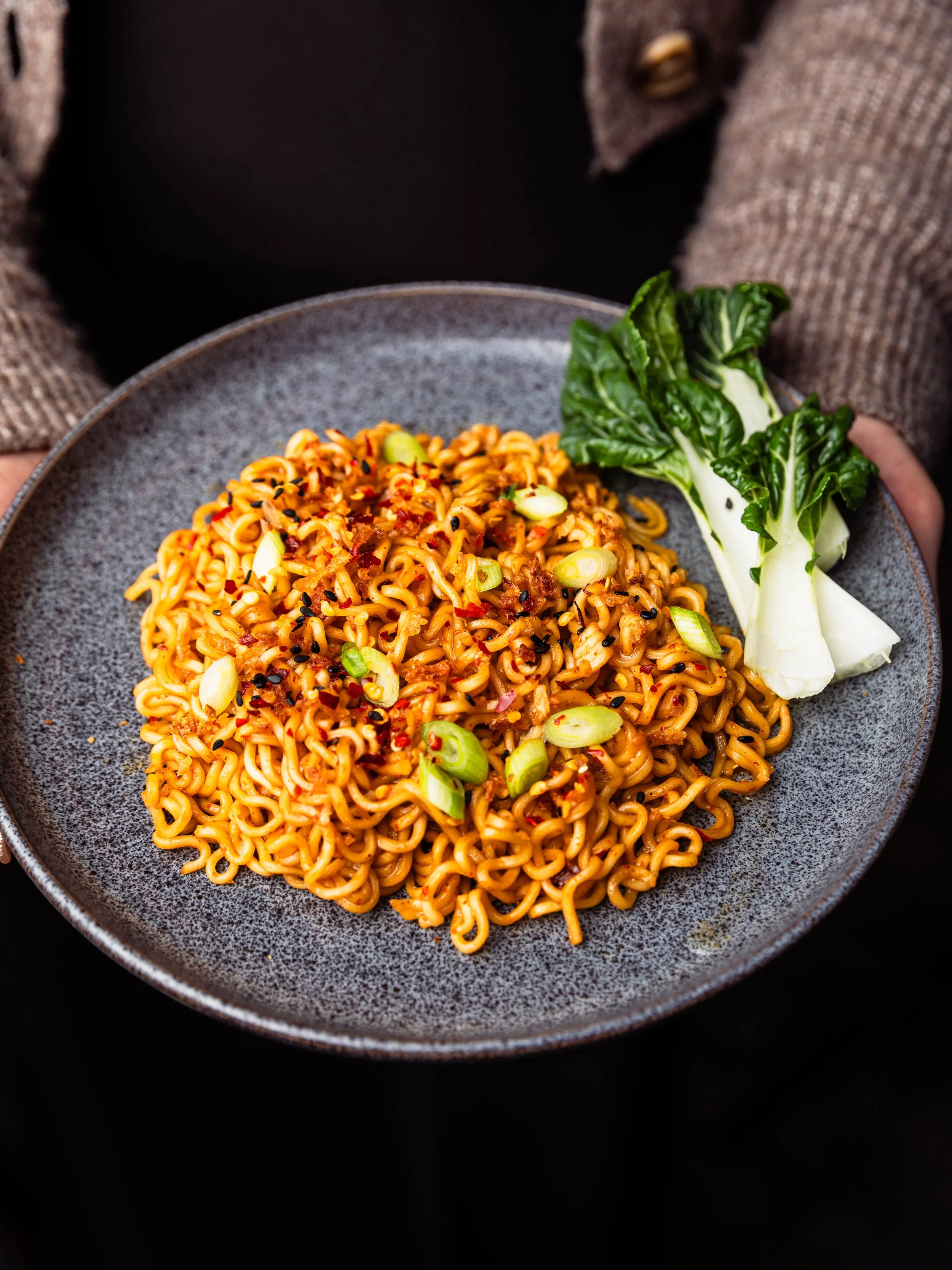 A plate of cooked instant ramen noodles topped with chopped green onions and red pepper flakes, served with a side of bok choy.