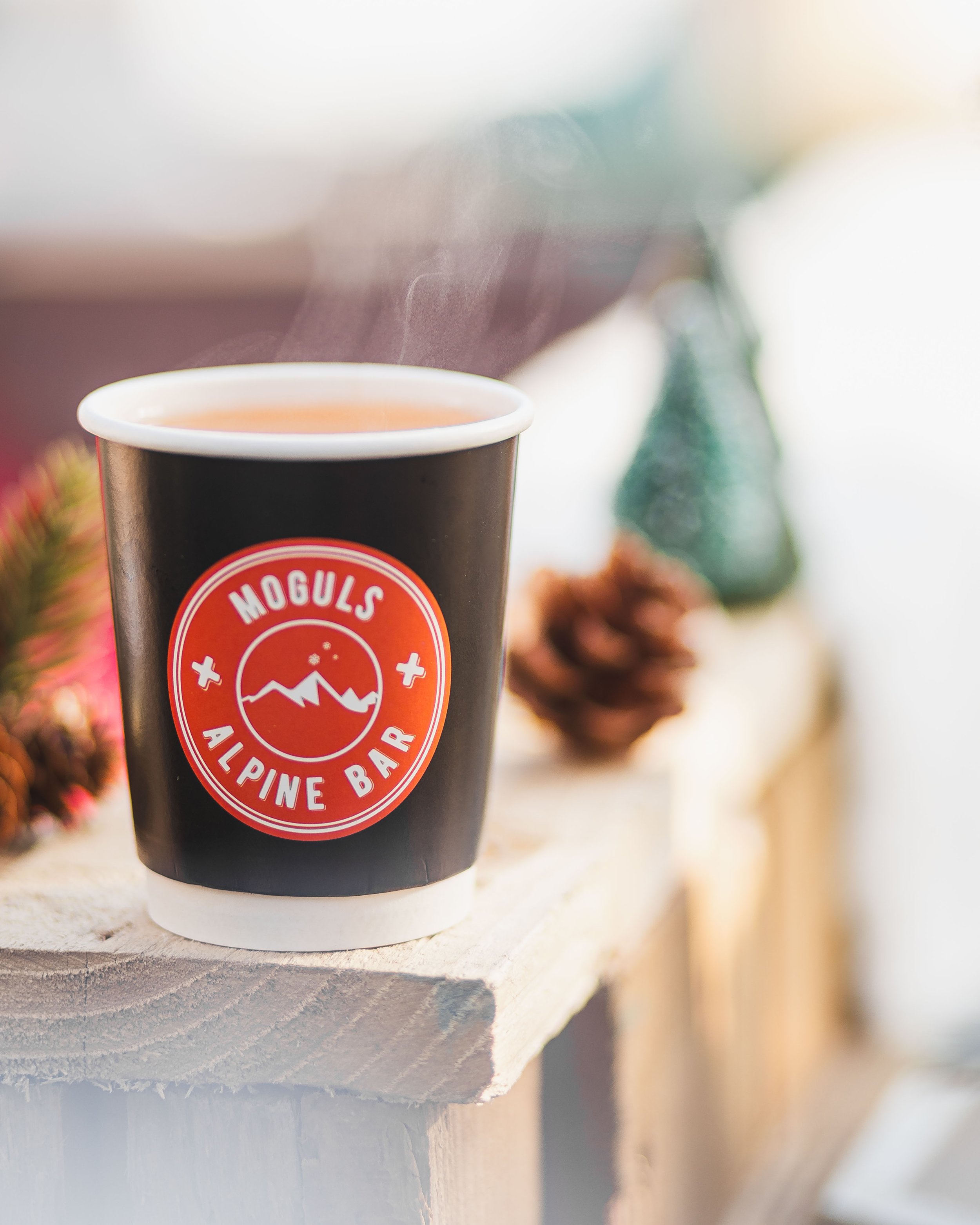 A steaming hot beverage in a black cup with a red and white logo for Moguls Alpine Bar, placed on a rustic wooden surface with out-of-focus pinecones and holiday decorations in the background.