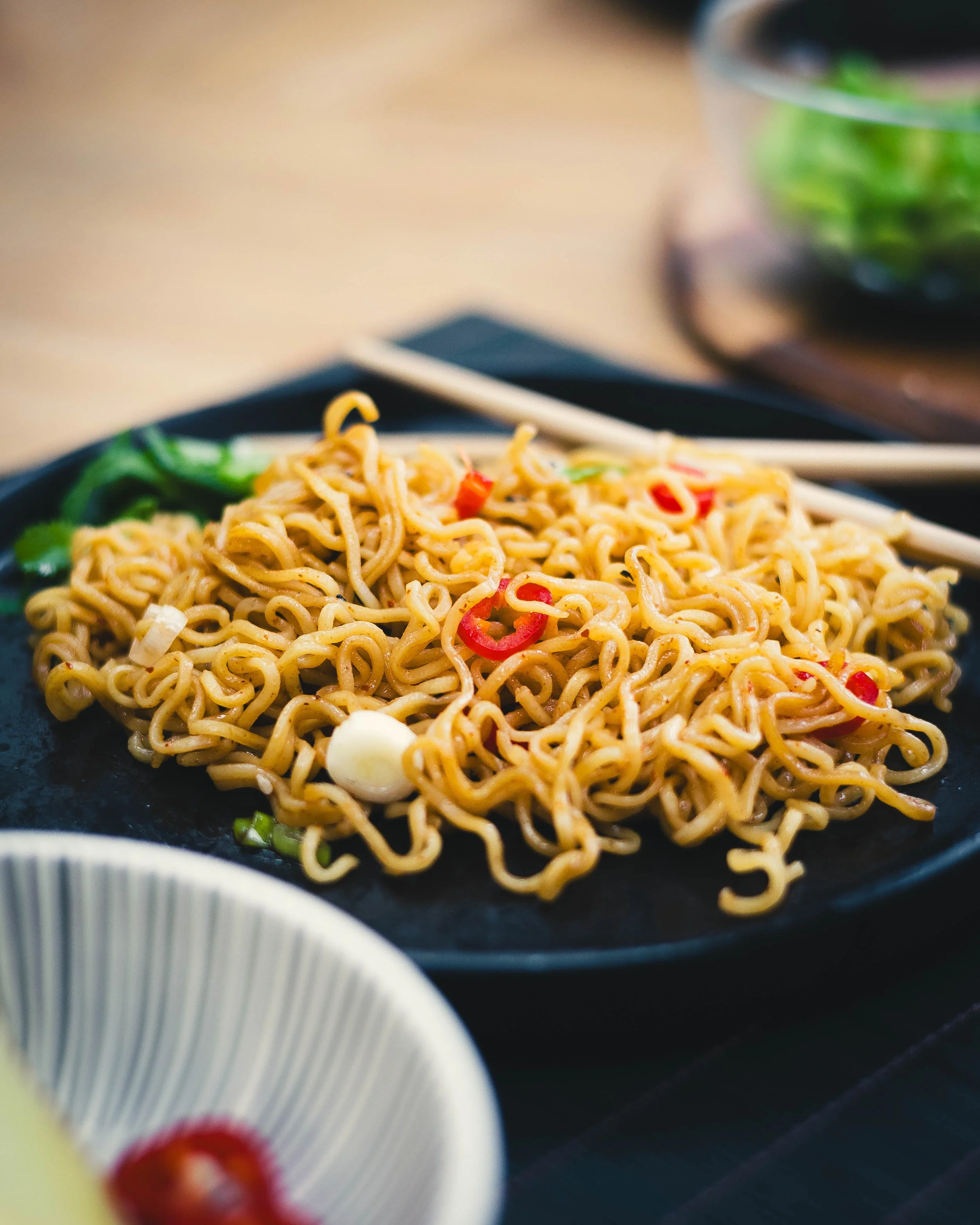 A black plate of ramen noodles garnished with sliced red chili peppers, green onions, and a boiled egg, set on a black tray with chopsticks.