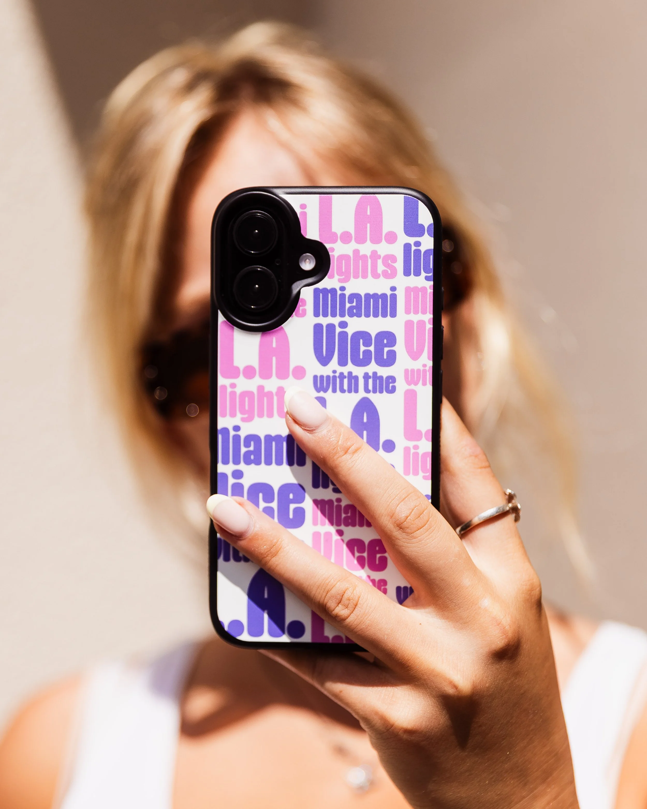 A woman with blonde hair and sunglasses taking a selfie with her phone, which has a colorful case with pink and purple text.