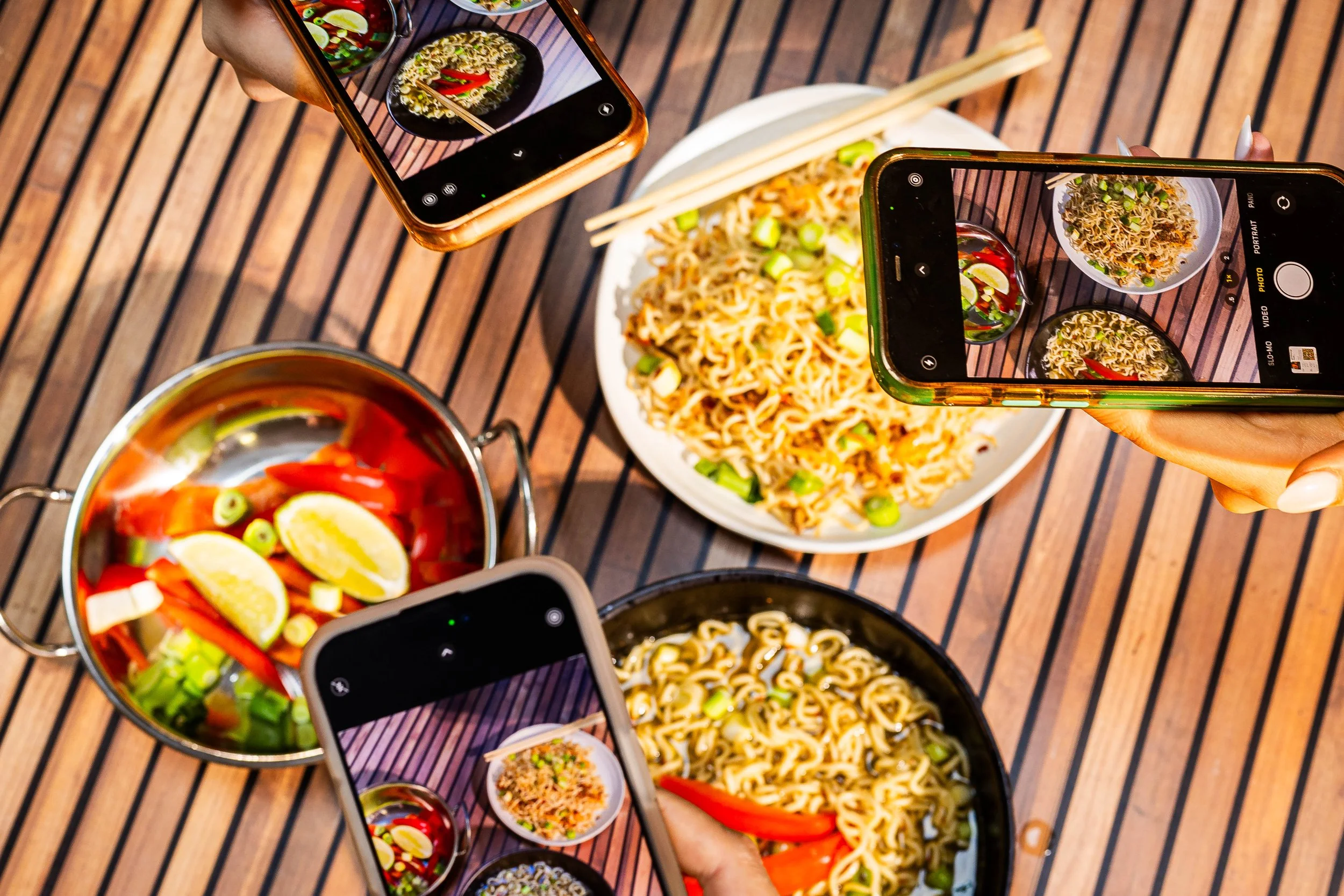 People taking photos of bowls of ramen noodles and a pot of sliced vegetables on a wooden table.