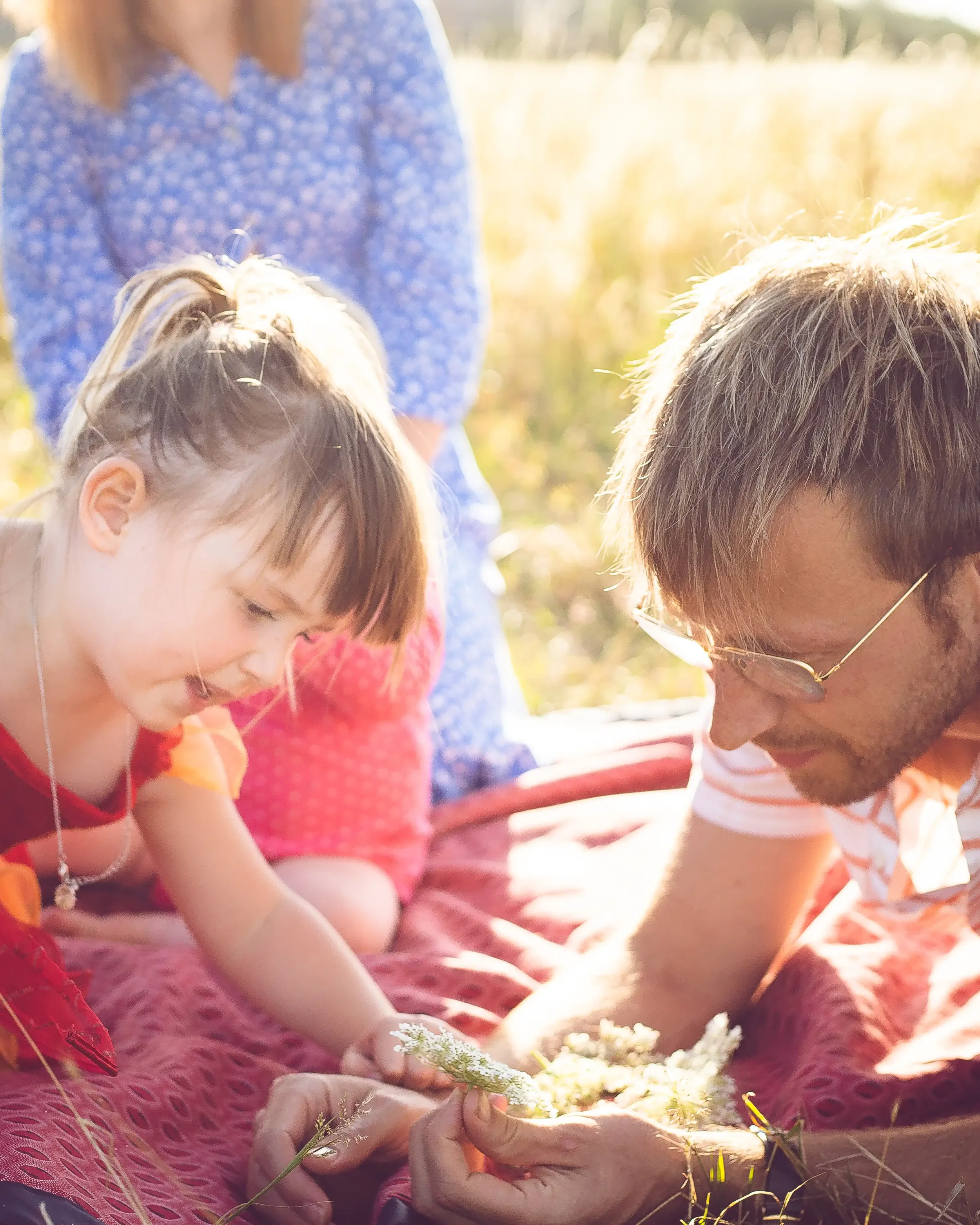A man and a young girl are lying on a blanket outdoors, closely examining flowers, with a woman standing behind them in a field during daytime.