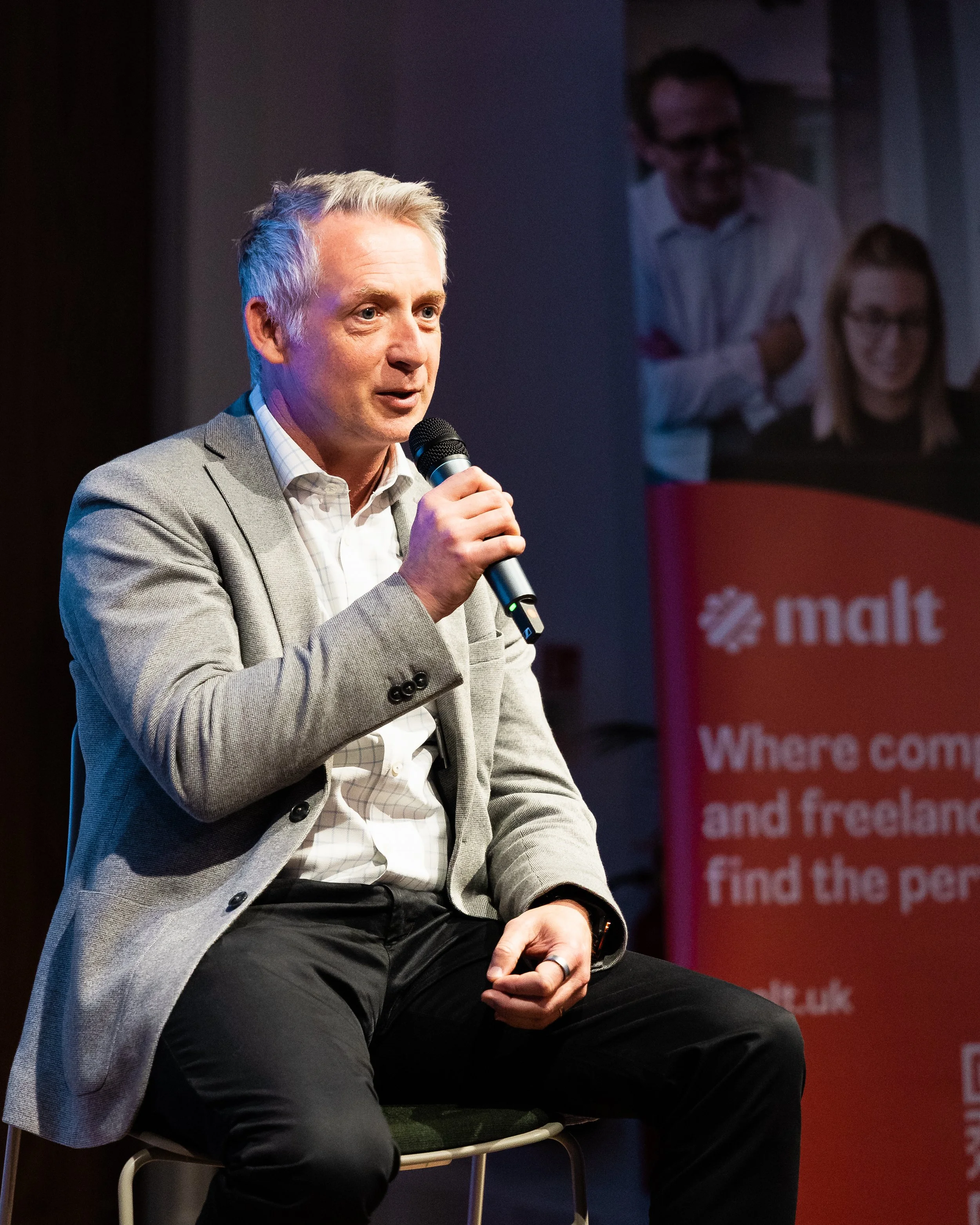 A man in a grey suit holding a microphone on stage at a business presentation by London Photographer Bobby Shoebotham