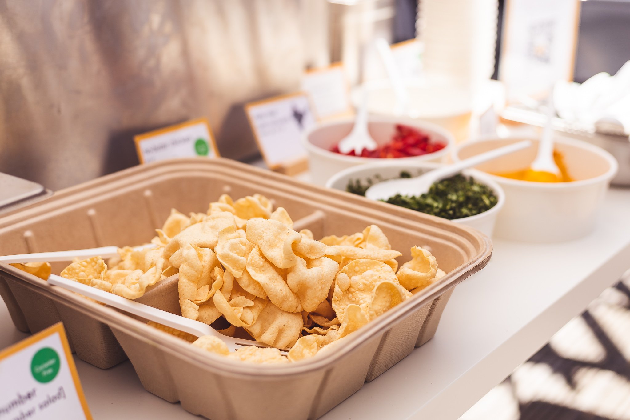 A cereal or snack bar station with a basket of crispy, fried snacks and bowls of toppings or condiments in the background.