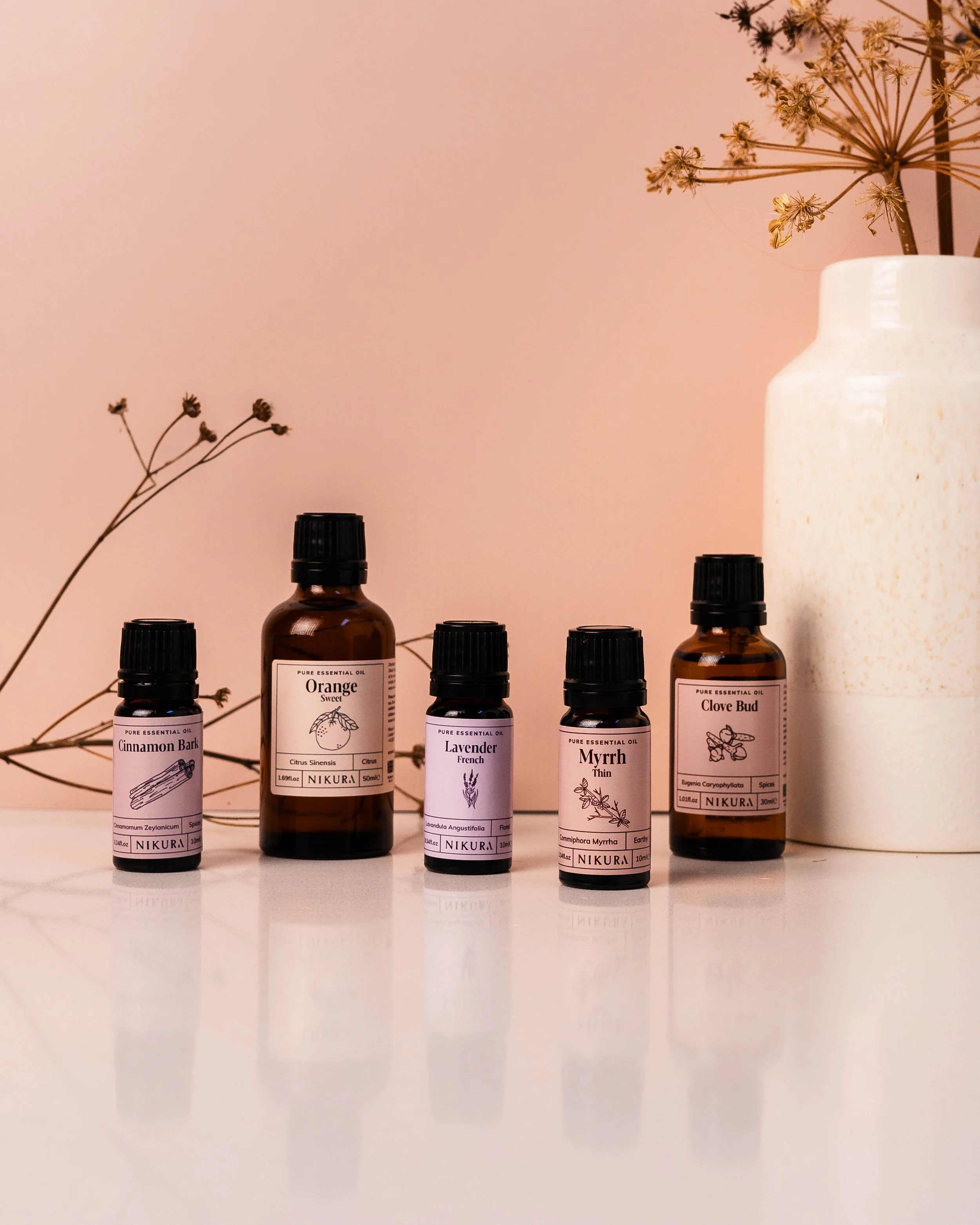 A selection of essential oils against a pink background next to a vase and some stylised twigs.
