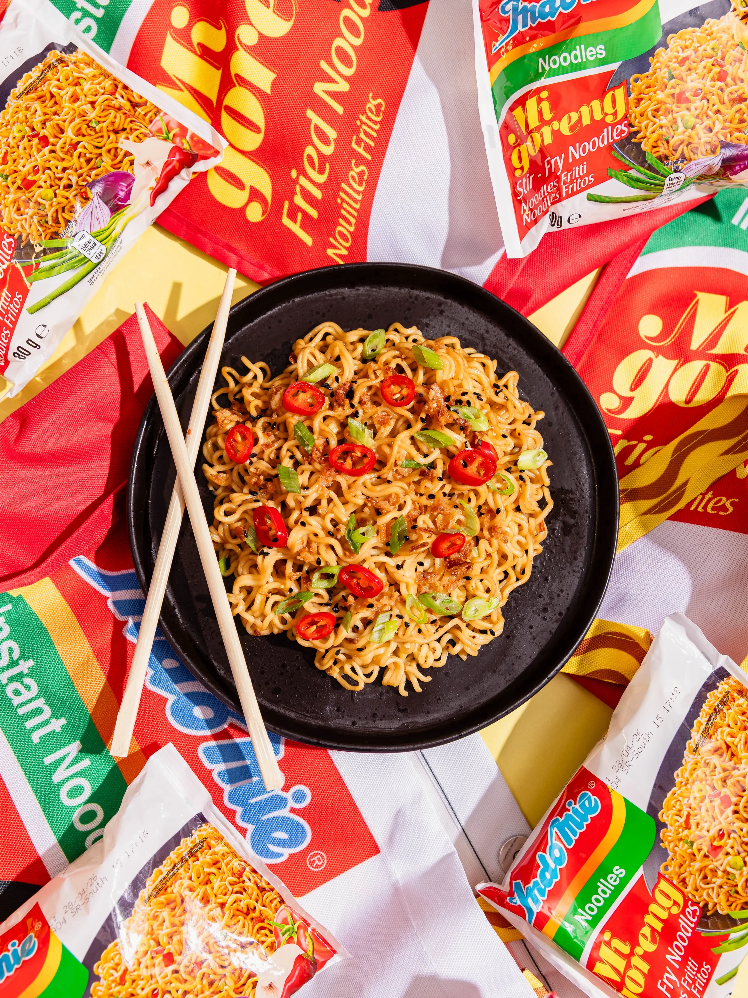 Plate of cooked instant noodles garnished with sliced red chili peppers and chopped green onions, surrounded by packs of instant noodles on a table.