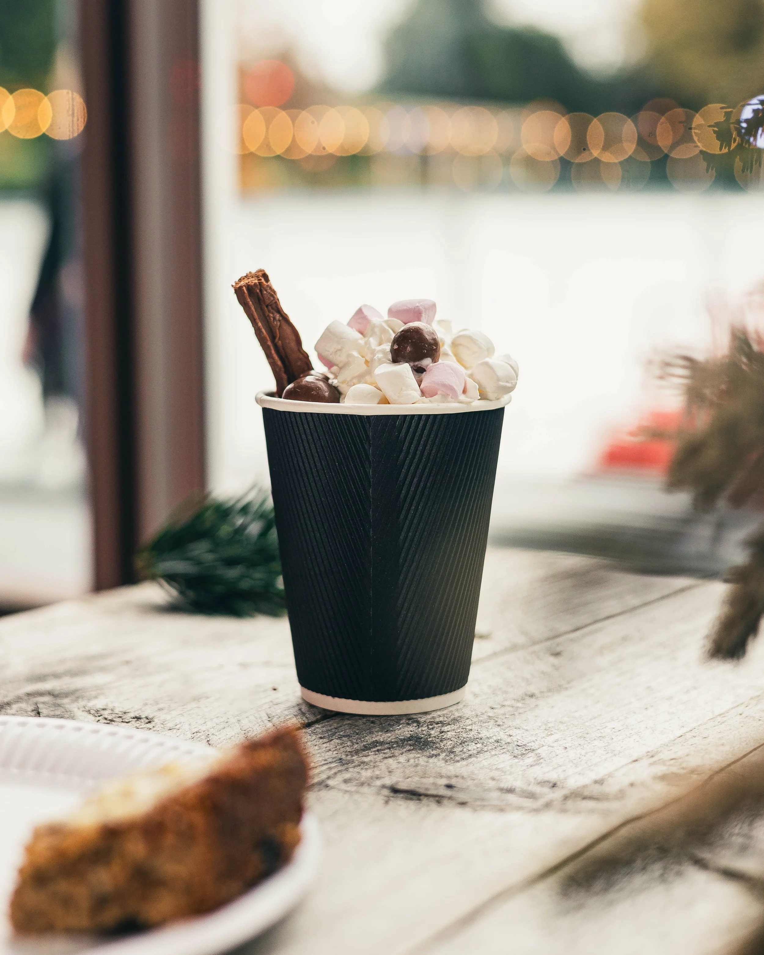 Hot chocolate topped with marshmallows, chocolate candies, and a chocolate wafer stick in a black paper cup, on a wooden table near a window with blurred background lights.