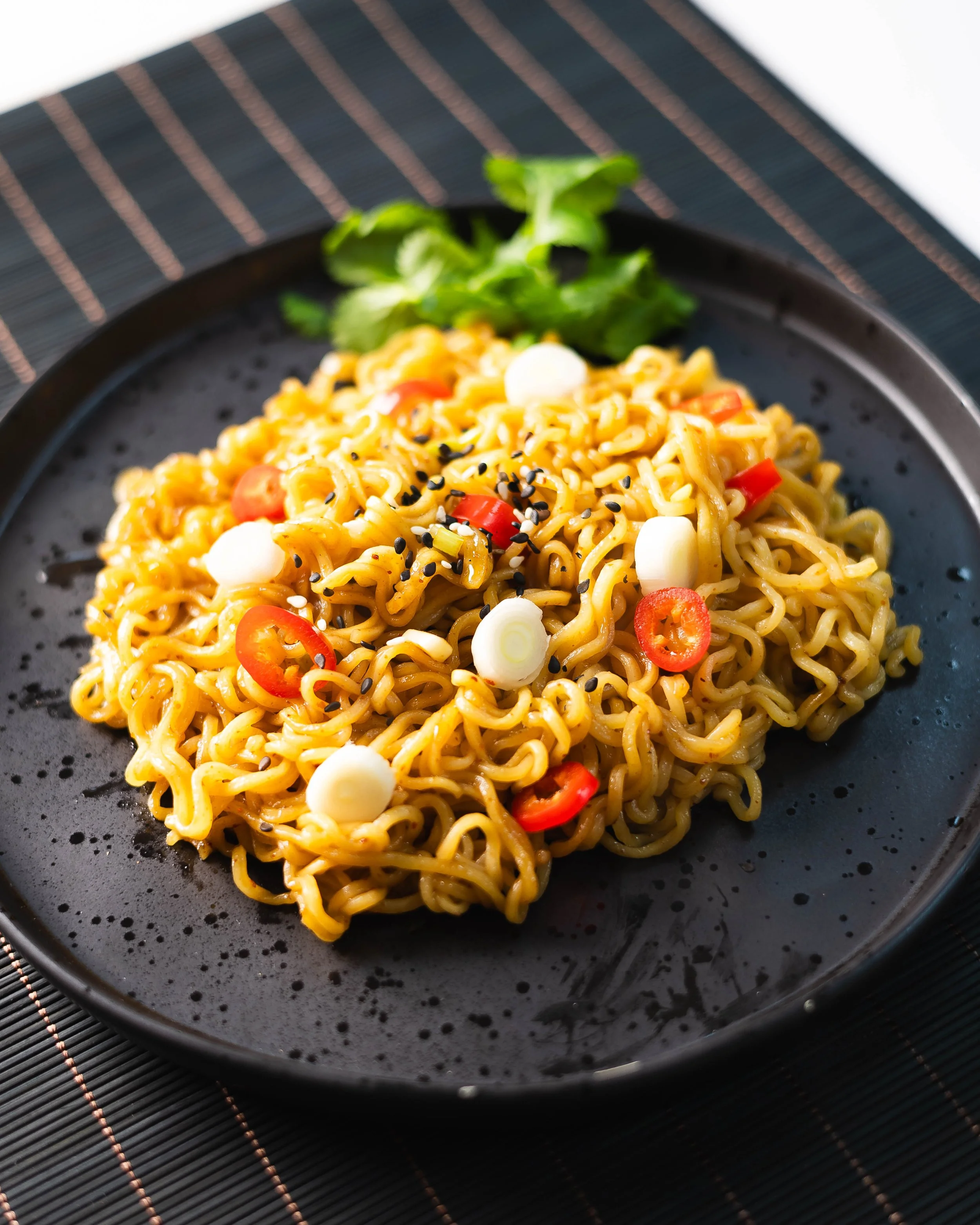 A black plate of spicy ramen noodles garnished with sliced red chili peppers, sliced green onions, black sesame seeds, and a sprig of cilantro, on a dark placemat.