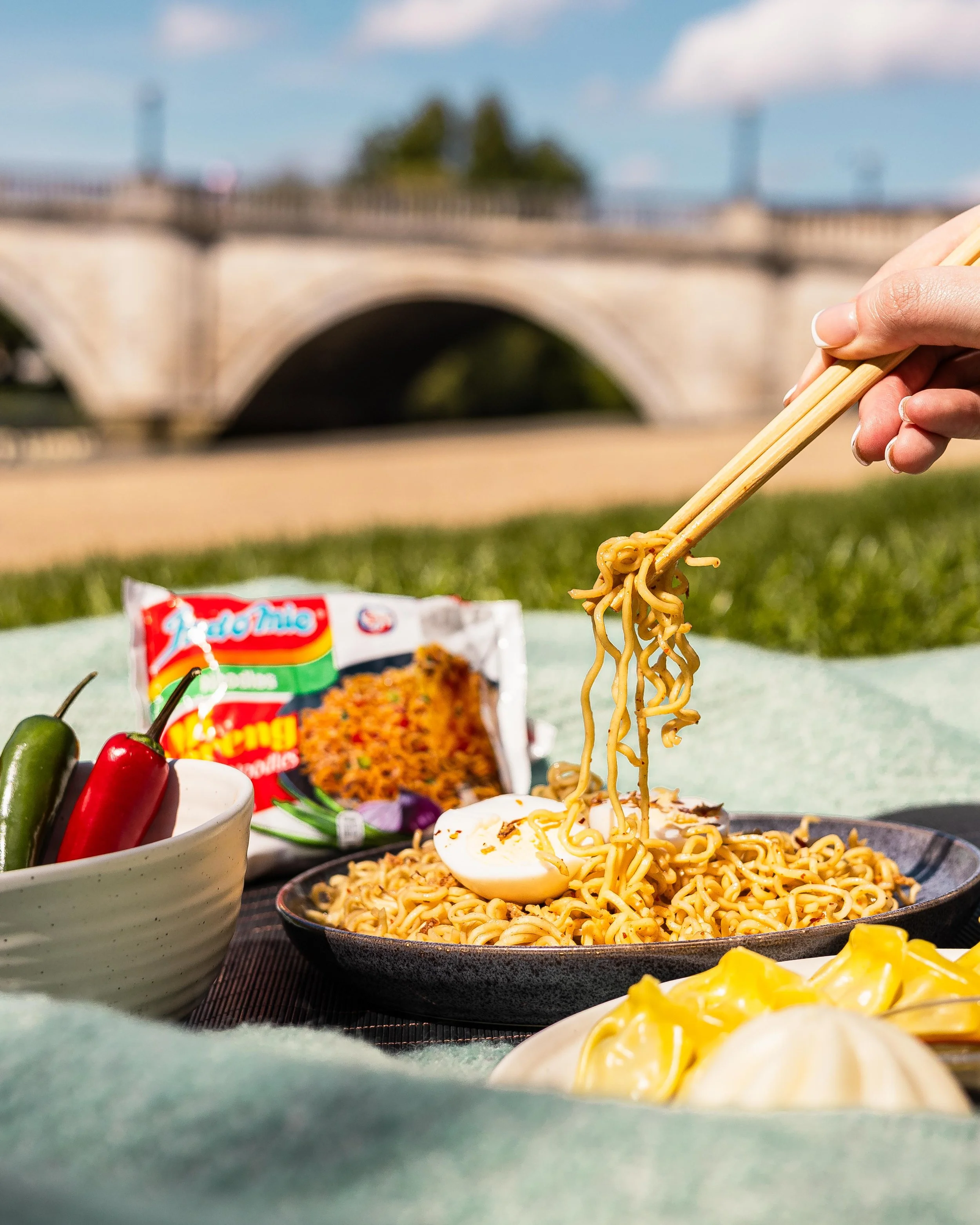 Hand holding chopsticks lifting noodles from a black bowl of ramen, with a soft boiled egg, on a picnic blanket outdoors with a bridge in the background.